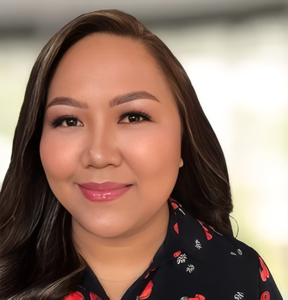 Close-up of a woman with long, dark hair, smiling, wearing a dark floral-patterned top, with a blurred background.