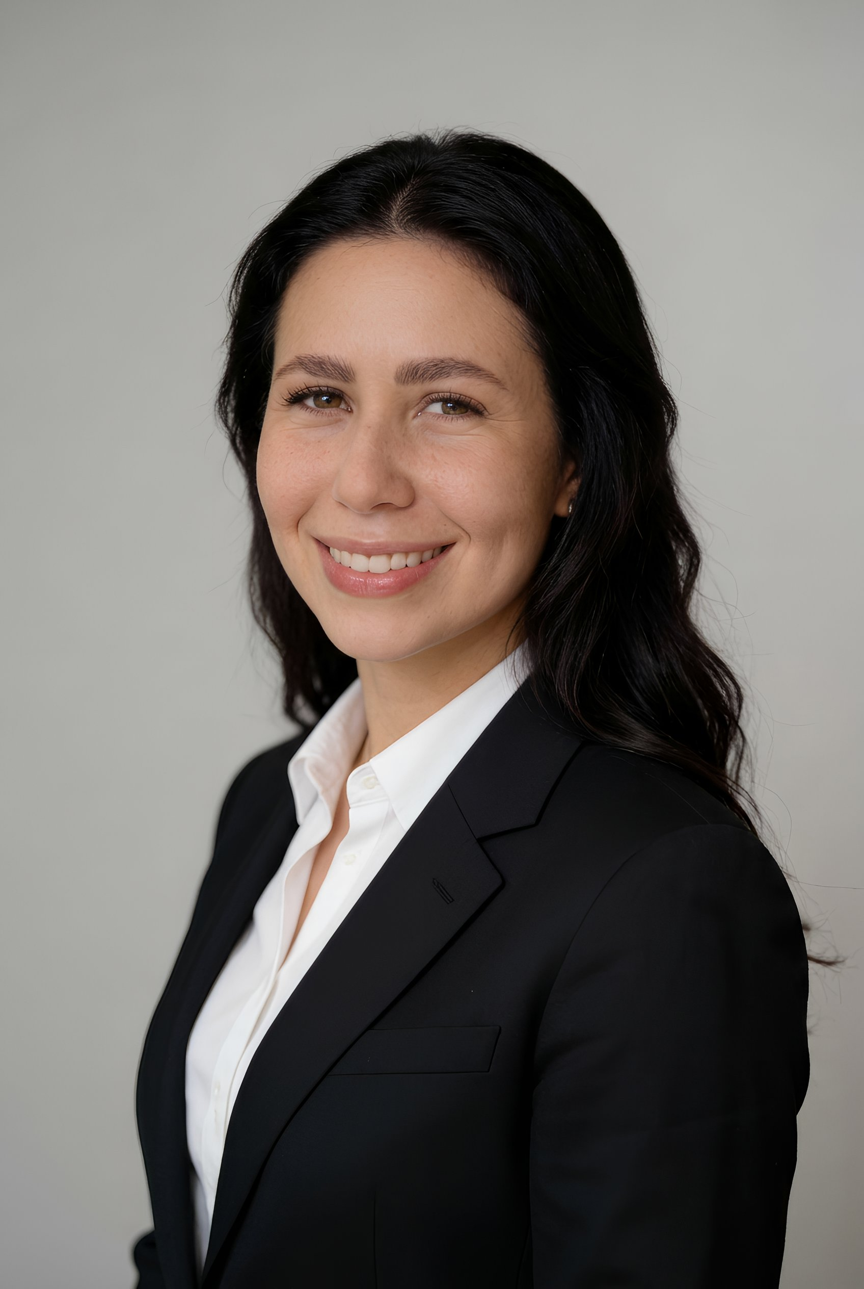 A professional woman with long dark hair, wearing a black blazer and white shirt, smiling at the camera against a plain background.