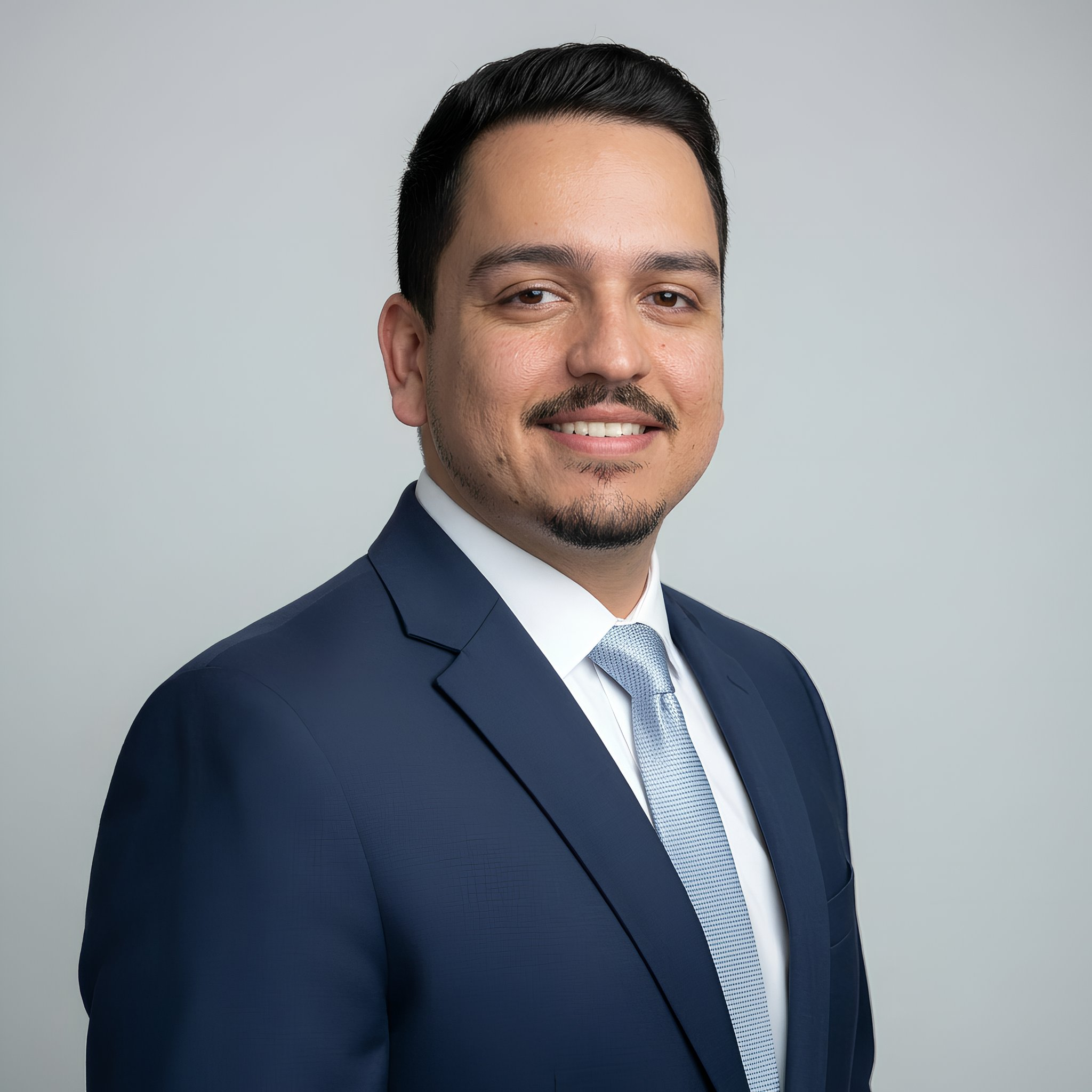 Professional headshot of a man with dark hair, a mustache, and goatee, wearing a navy blue suit, white shirt, and light blue tie, smiling against a plain light gray background.
