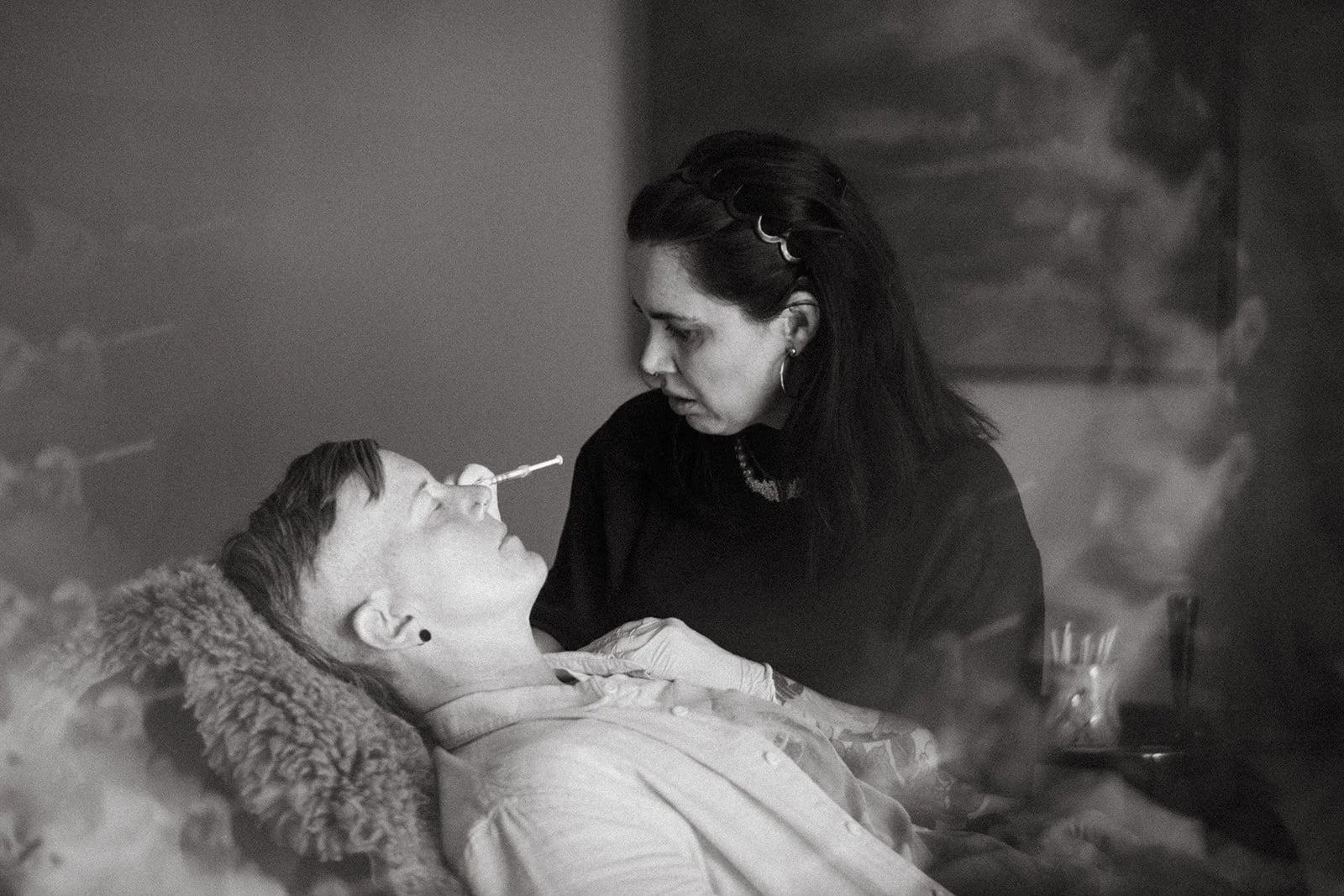 A woman with dark hair, earrings, and a necklace leans over a person lying in bed, holding their chin. The person in bed has short hair, earrings, and is having a tox treatment done. The scene is calm and intimate.