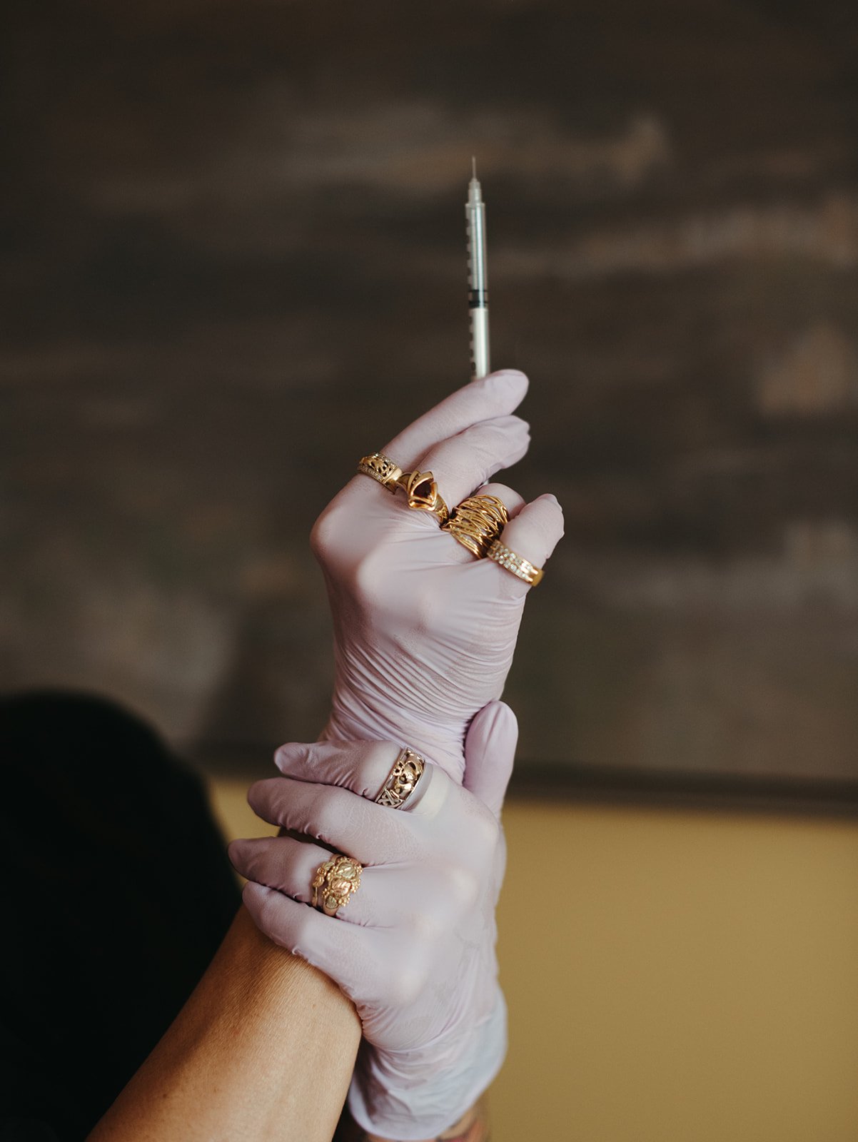 A hand wearing multiple gold rings and a white glove holding a syringe with a needle pointing upwards.