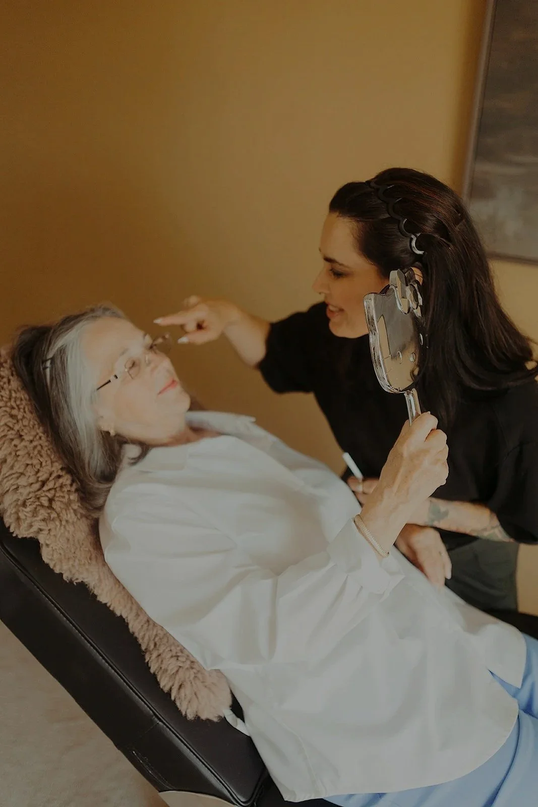 A woman with dark hair and tattooed arm gently touching a senior woman with gray hair and glasses, lying in a chair, while holding a mirror.