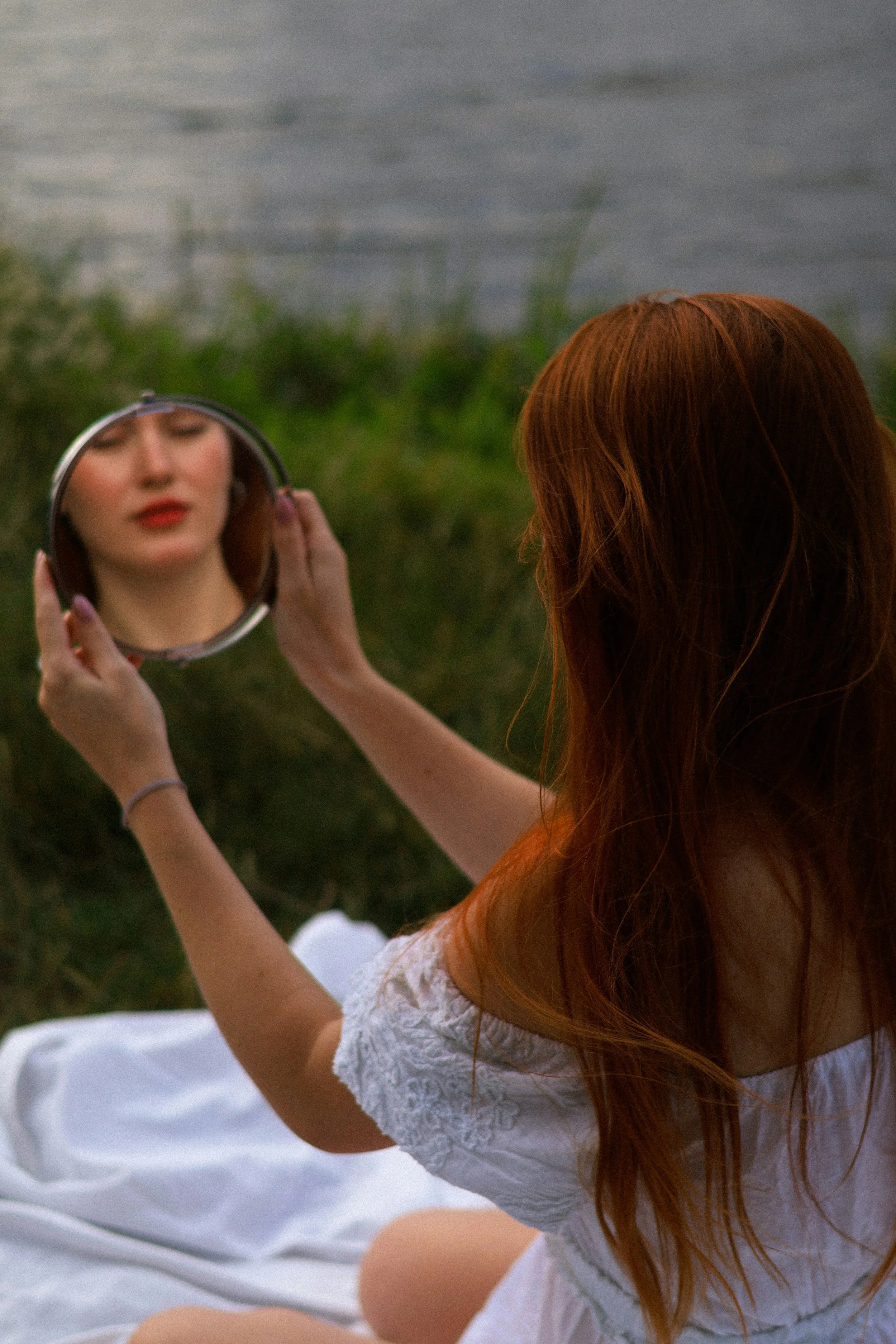 A woman with long red hair sitting outdoors on a white blanket, looking into a mirror and adjusting her makeup, with a lake and greenery in the background.