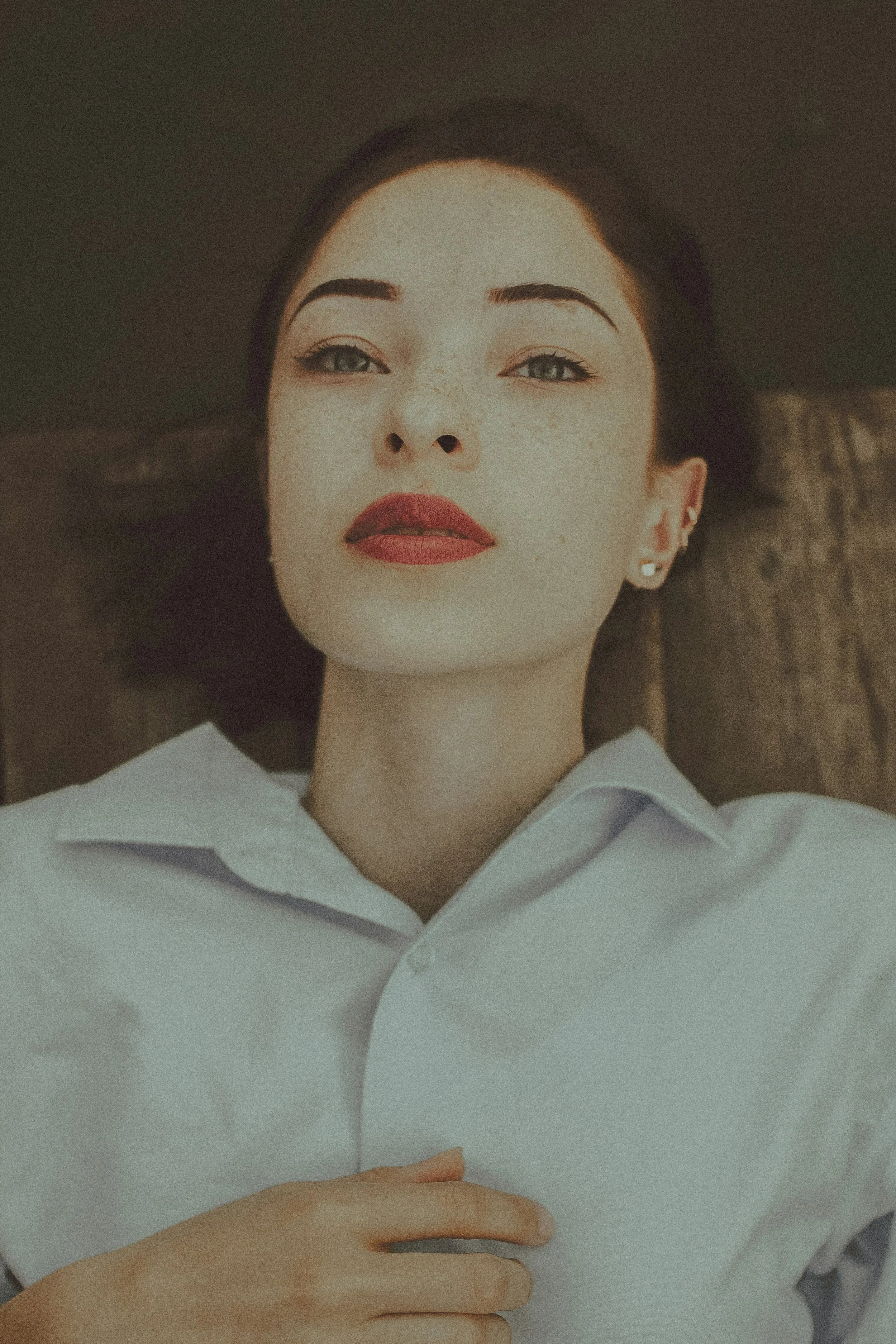 A woman lying down with her head on a surface, wearing a white shirt, with makeup and earrings, looking directly at the camera.