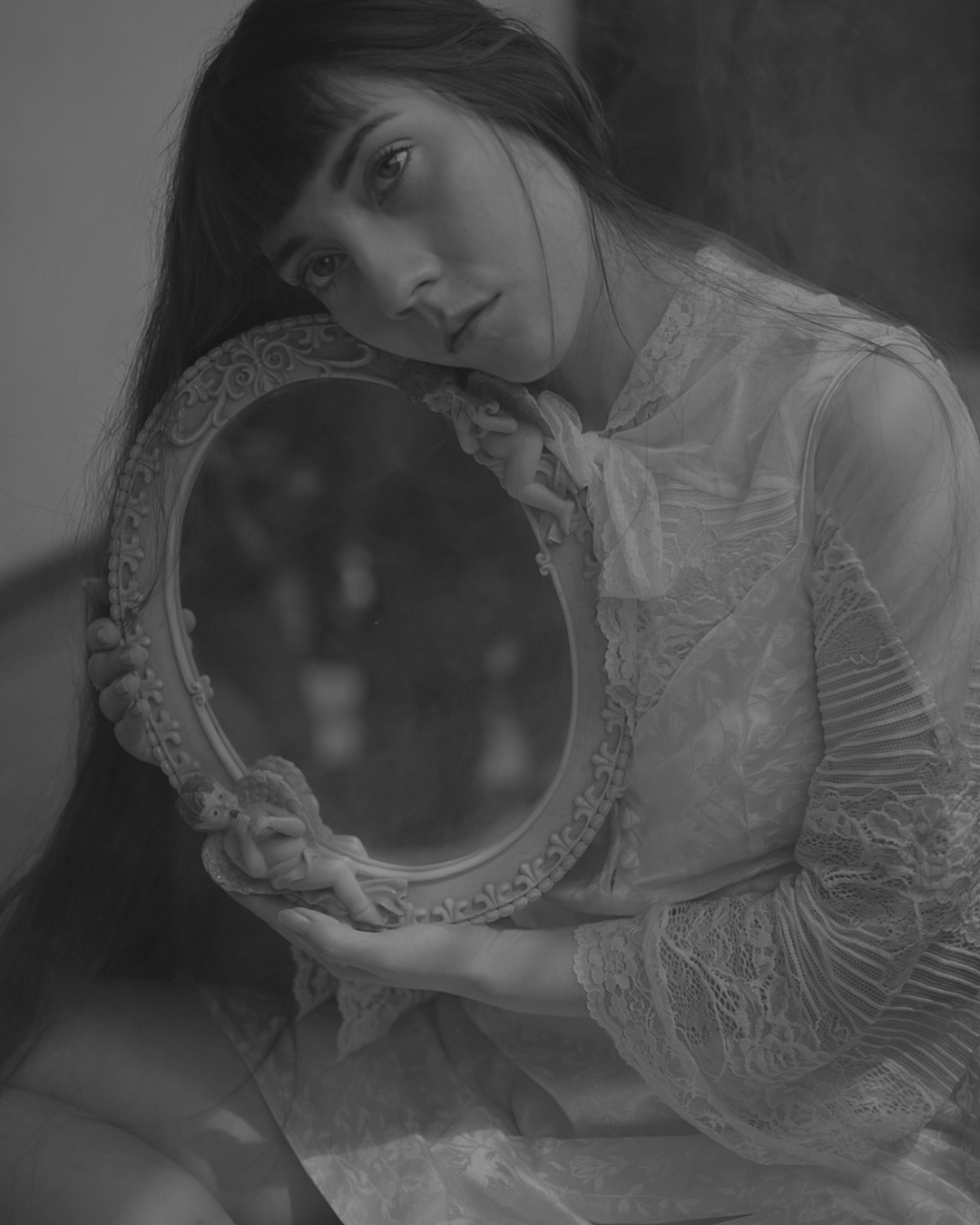 A woman with long hair and bangs, wearing a lace dress, holding a decorative mirror with a cherub design on the frame, looking into it.