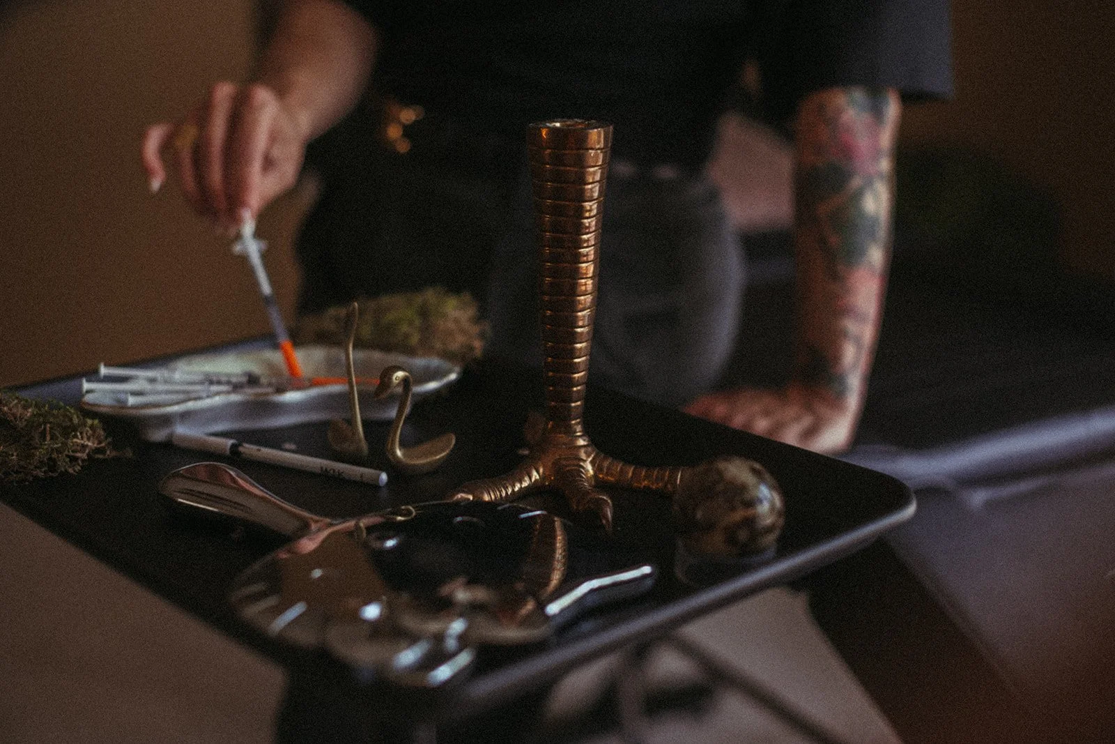 A person with tattooed arms is seen near a black table with various objects, including a metallic sculpture of a bird, a large copper-colored spiral object, and some tox treatment syringes.