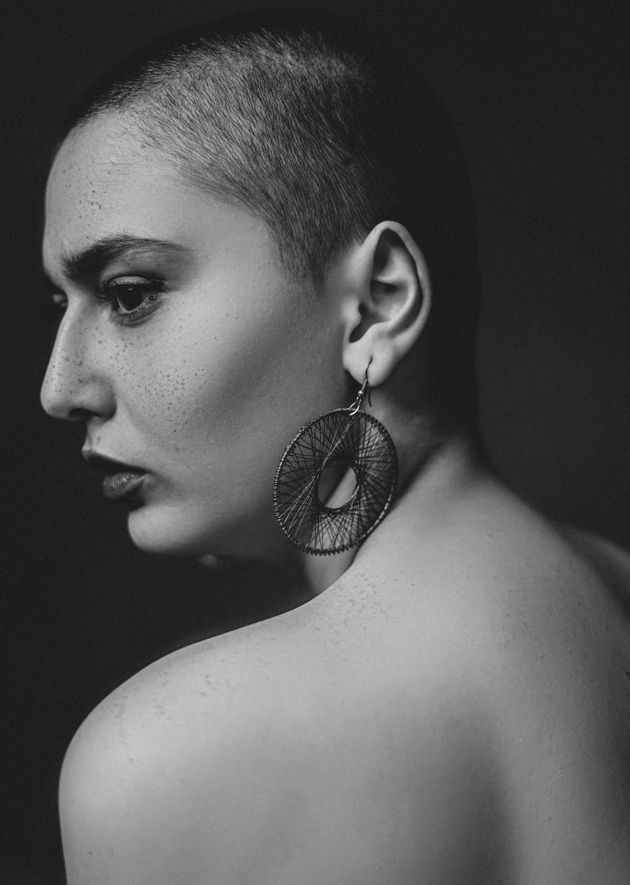 A black-and-white portrait of a woman with a buzz cut hairstyle, wearing a large circular earring, looking to her left, with smooth skin and freckles visible on her face and shoulder.