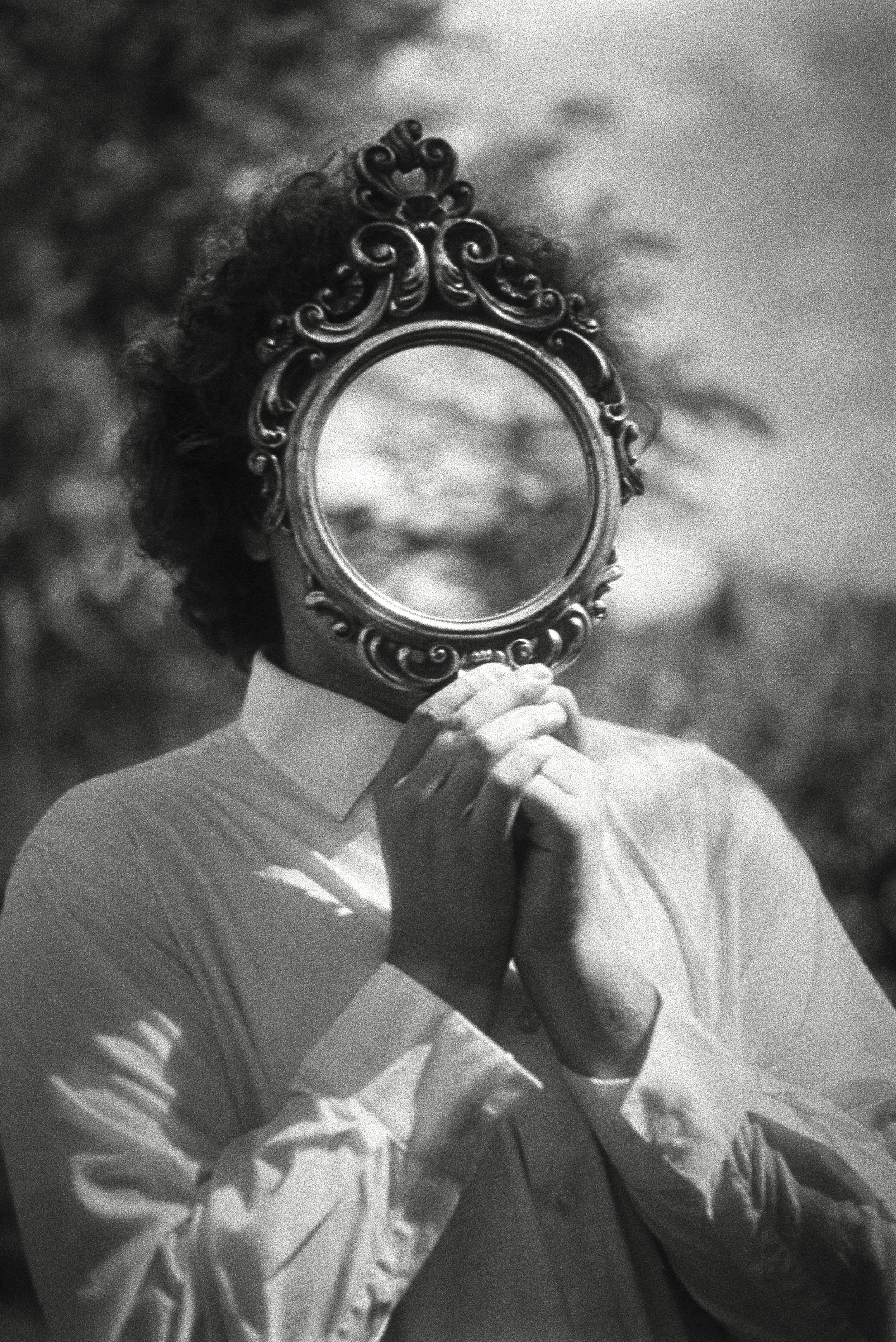 A person in a collared shirt holding a decorative round mirror in front of their face, obscuring it. The mirror has an ornate, intricate frame. The person appears to be outdoors, with blurry trees or foliage in the background.