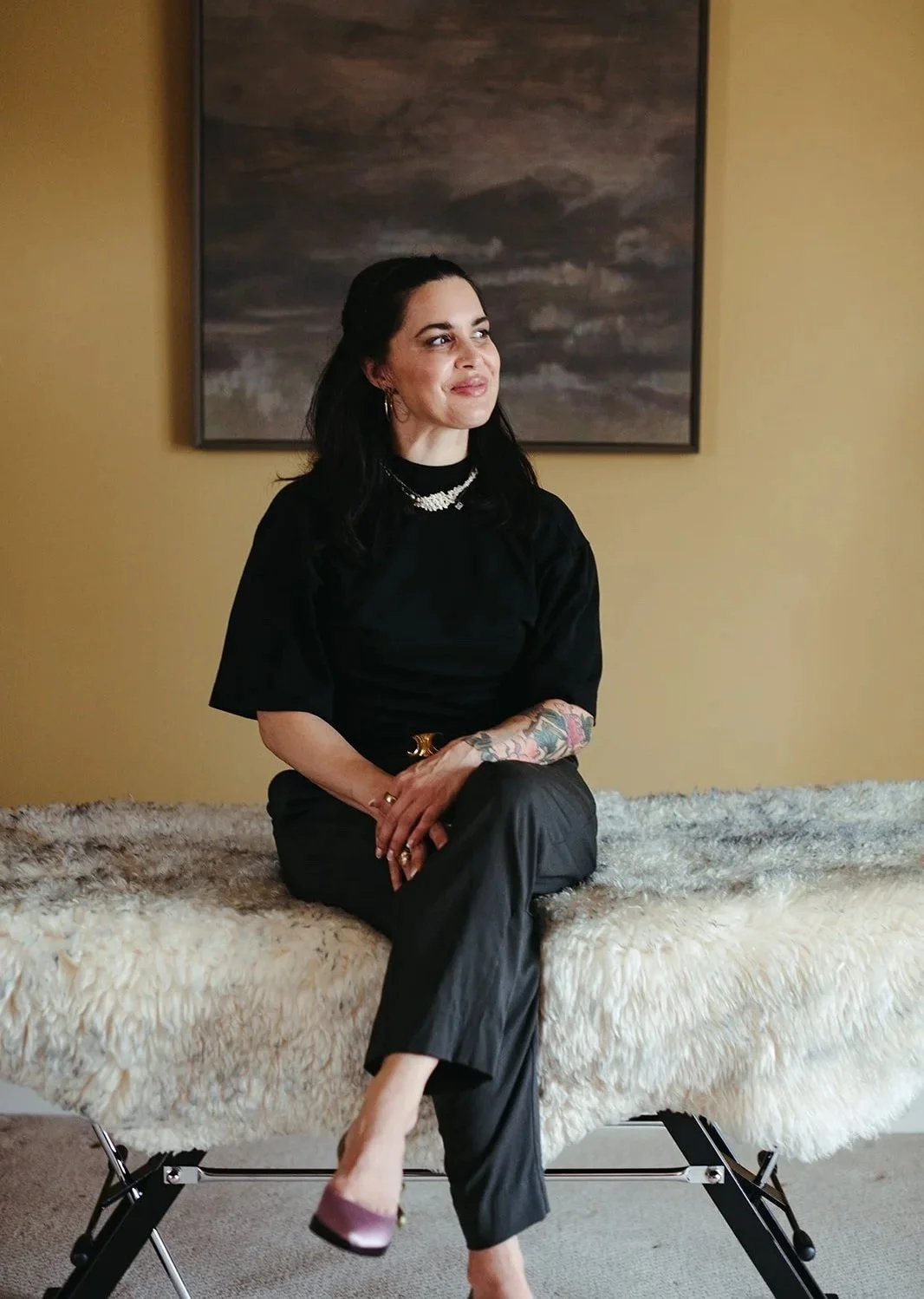A woman with dark hair and tattoos on her arm, sitting on a plush cream-colored surface with her legs crossed, wearing a black top and black pants, in front of a framed painting of a cloudy sky on a yellow wall.