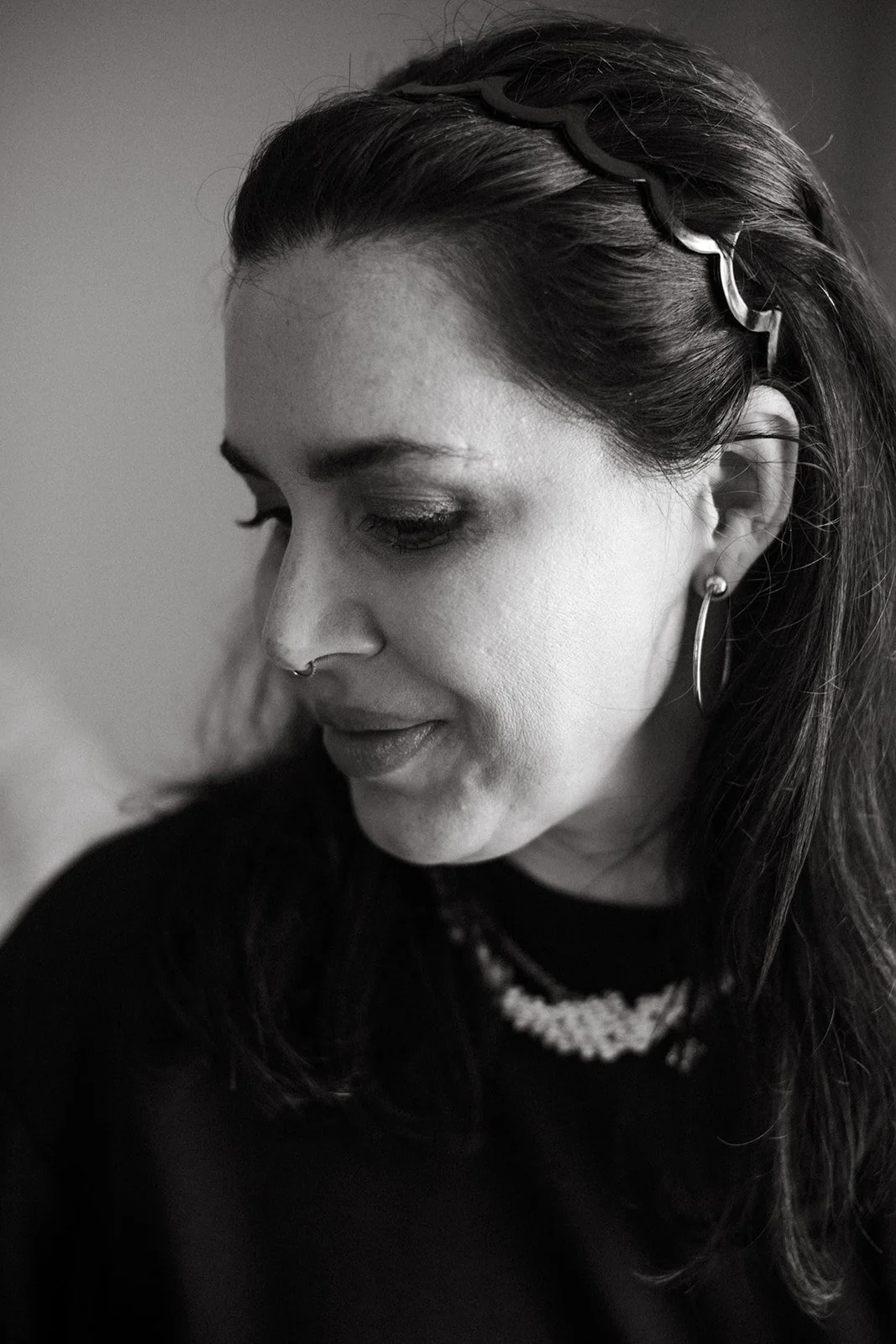 Black and white side profile of a woman with dark hair, wearing earrings, a nose ring, and a beaded necklace, looking downward.