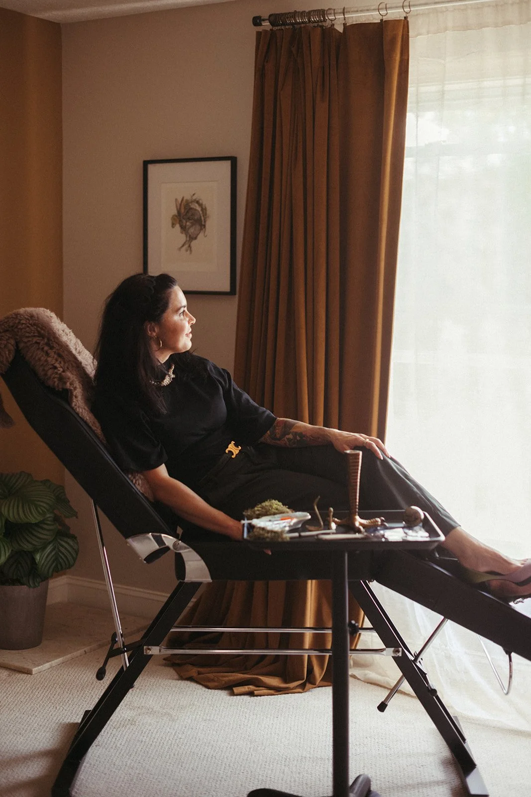 Woman sitting on a reclining chair near a window with golden curtains, looking out thoughtfully.
