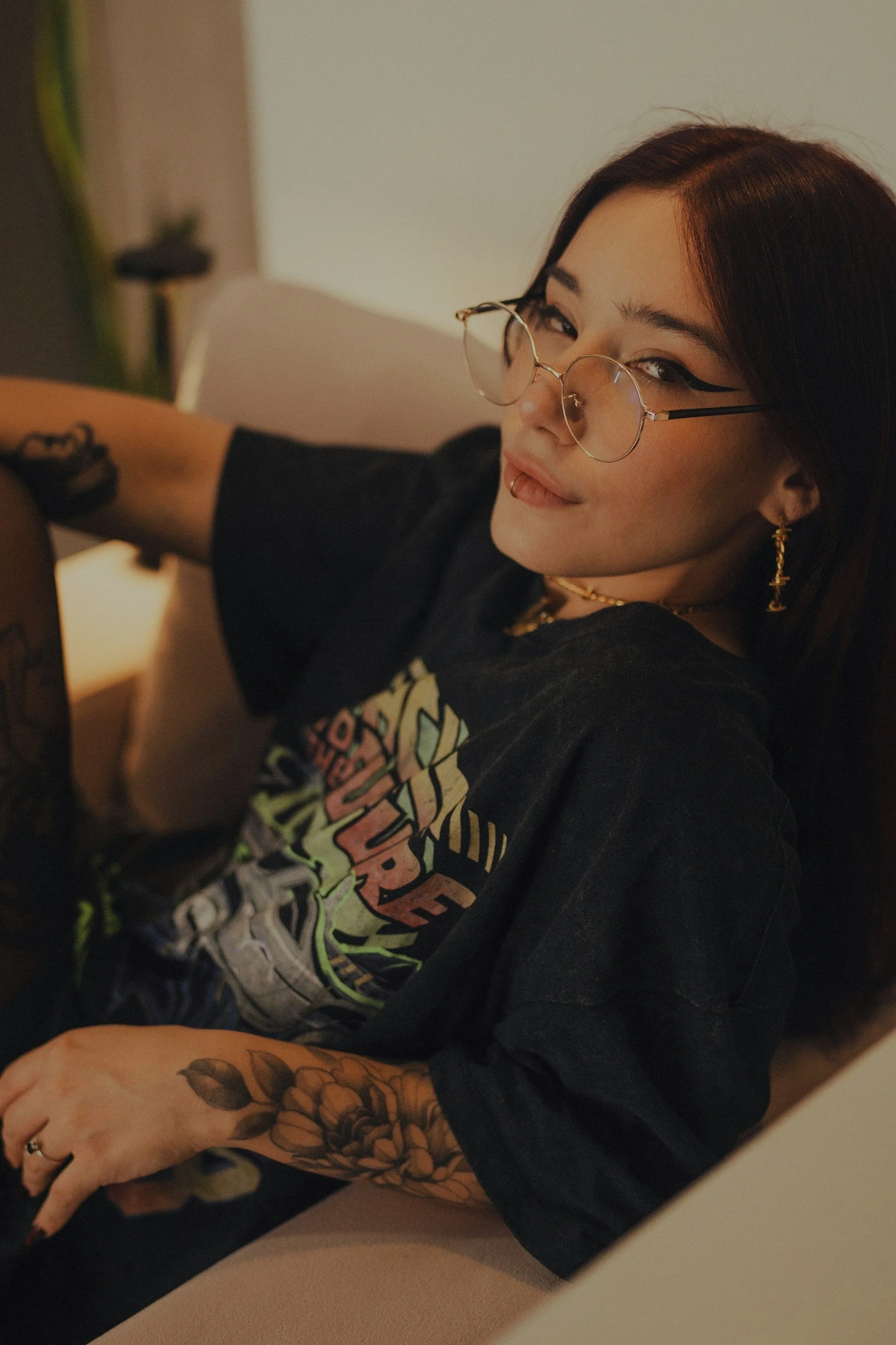 A woman with glasses and earrings sitting on a beige couch, wearing a black graphic T-shirt with colorful writing and tattoos on her arm.