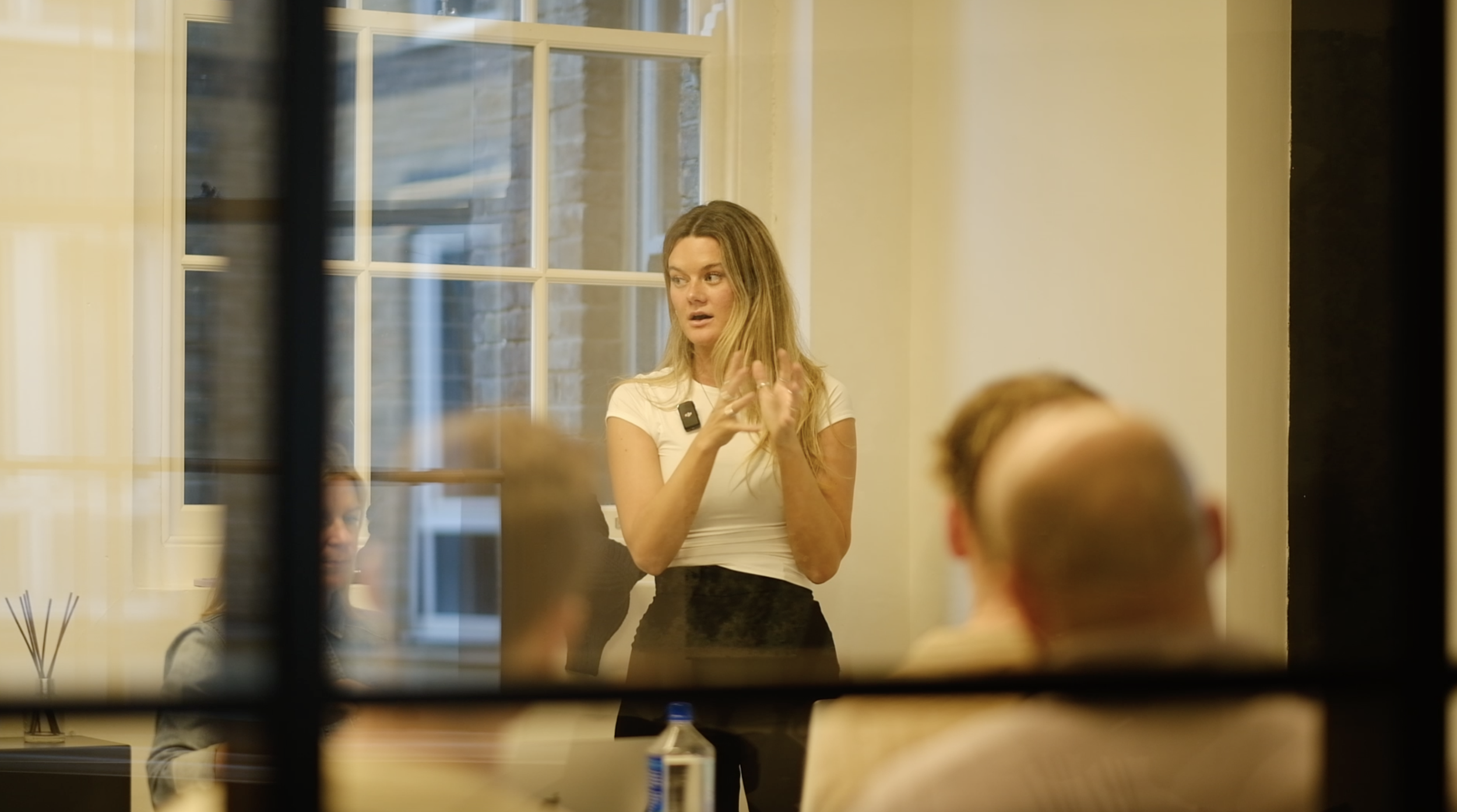 A woman standing and speaking, gesturing with her hands during a presentation in a room full of seated people, seen through a window or glass partition.