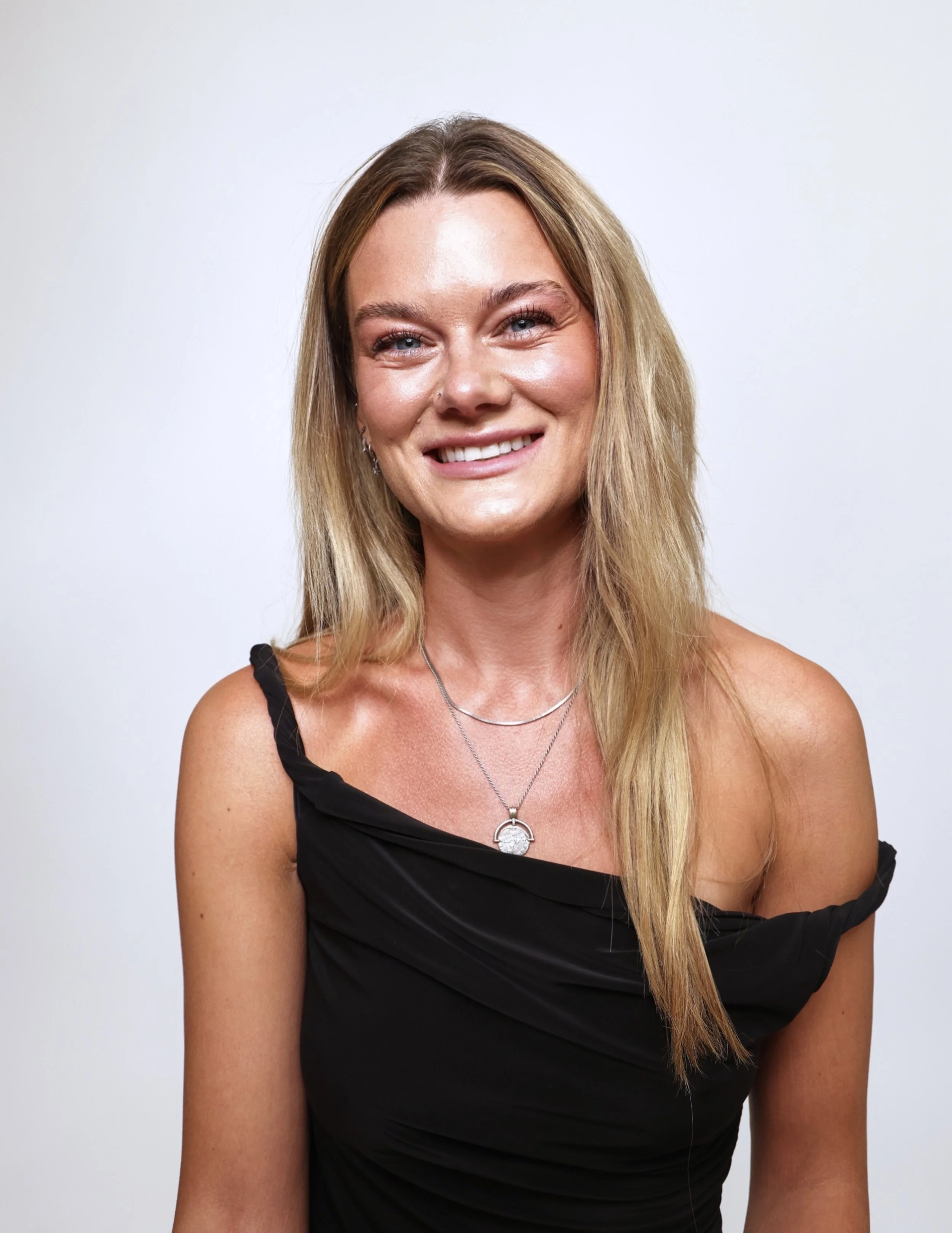 Portrait of a smiling woman with blonde hair wearing a black off-shoulder dress and silver jewelry against a plain white background.