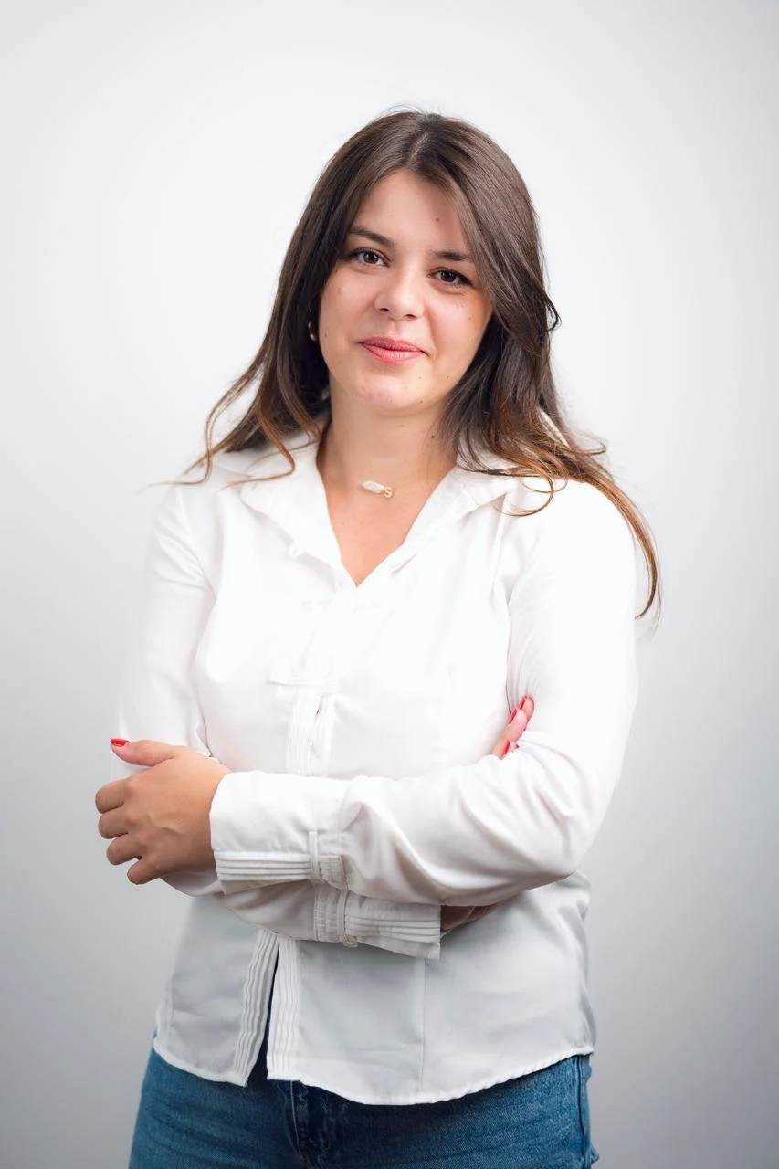 A woman with brown hair wearing a white blouse and blue jeans, standing against a plain white background with arms crossed.