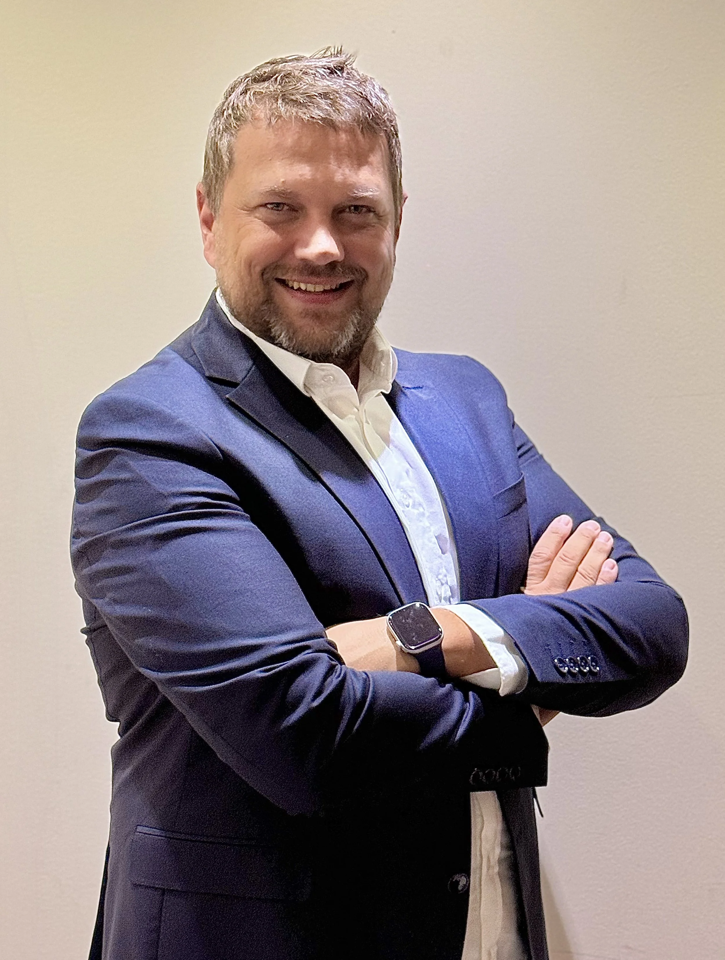 A man in a dark blue suit with a white shirt, smiling with arms crossed, standing against a plain beige wall.