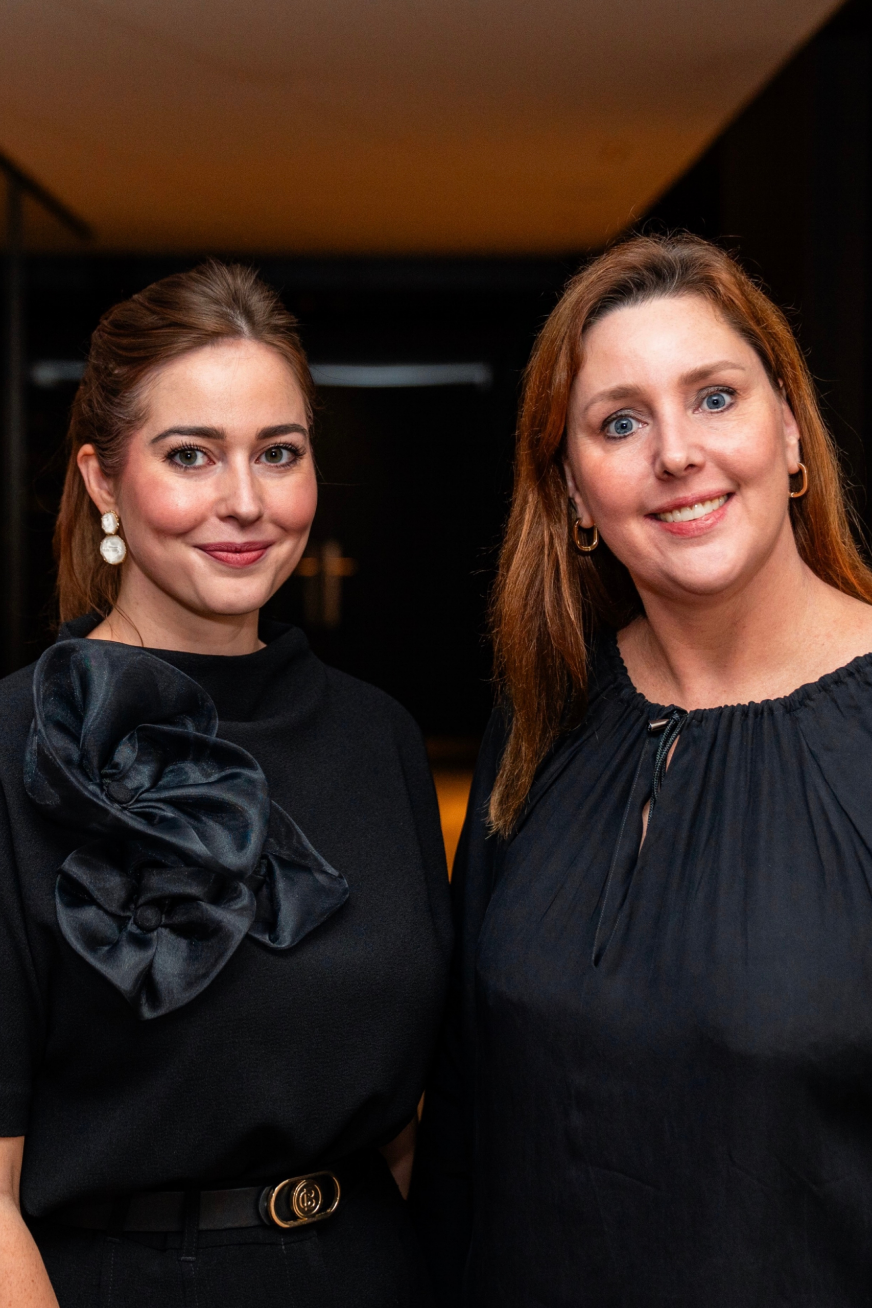 Two women smiling in an indoor setting, wearing dark outfits.