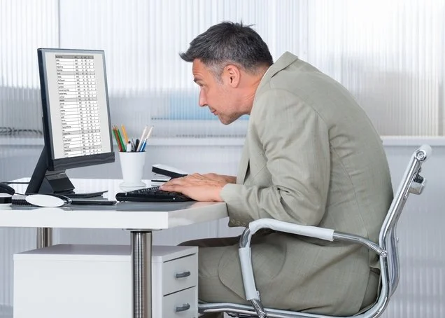 mala Ergonomía. Hombre en oficina revisando una hoja de cálculo en una computadora, sentado en una silla moderna, con objetos de oficina en el escritorio.