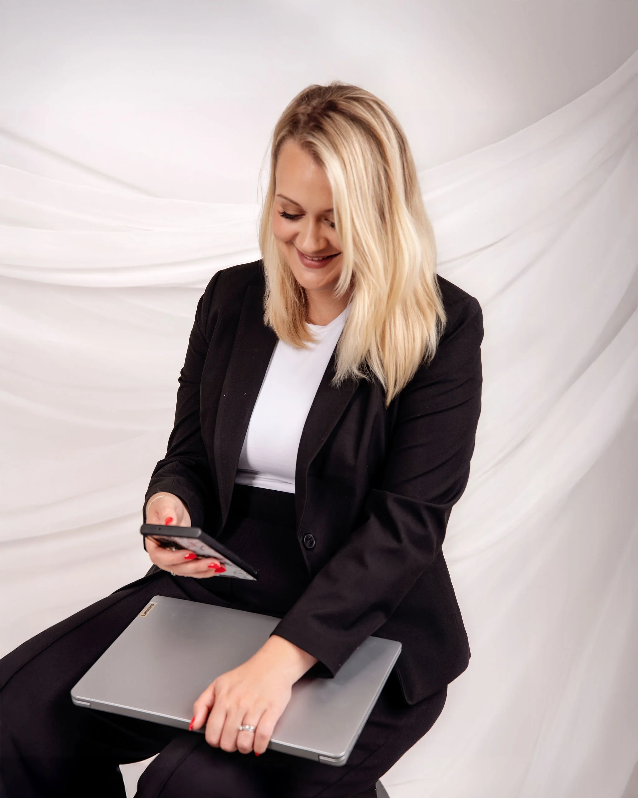 A woman sitting down, holding a smartphone, with a laptop resting on her lap, smiling and looking at her phone in front of a plain white background.