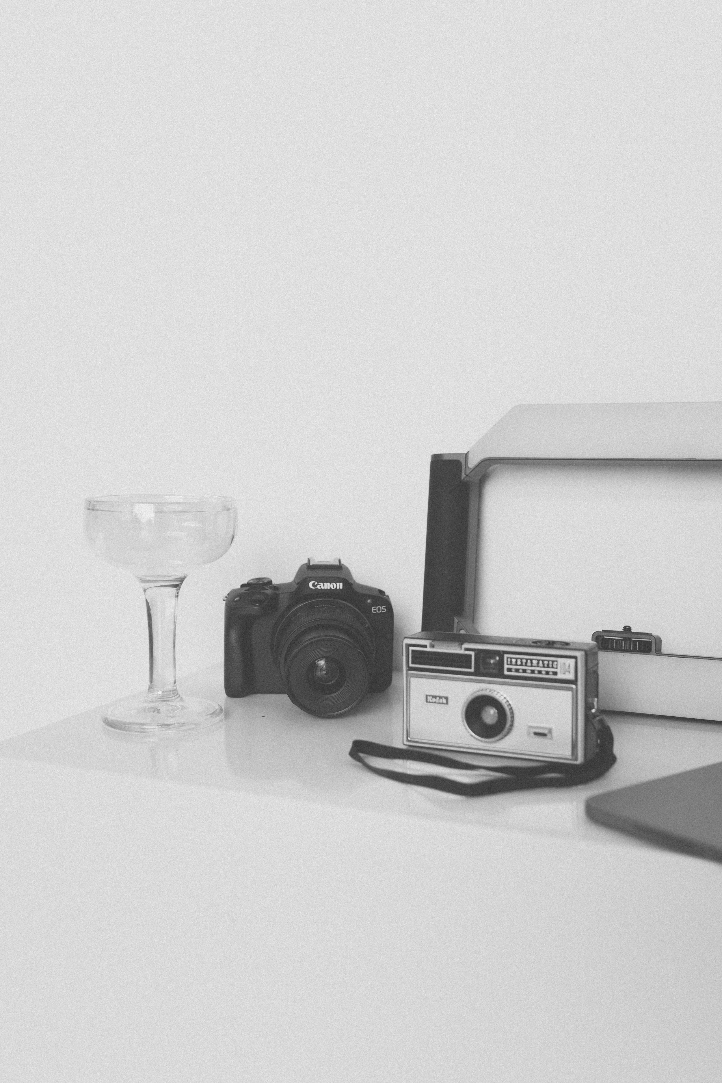 Black camera, vintage camera with strap, empty cocktail glass, photo box, and part of a laptop on a white surface.