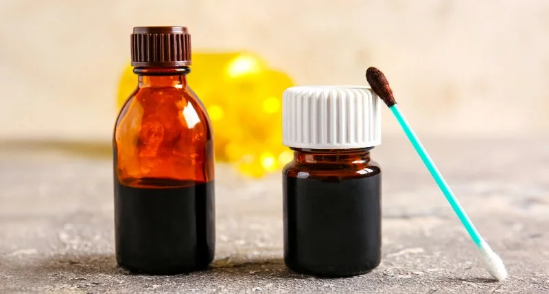 Two small amber glass bottles with white and brown caps, next to a cotton swab with a dark substance on it, on a textured gray surface with yellow objects blurred in the background.