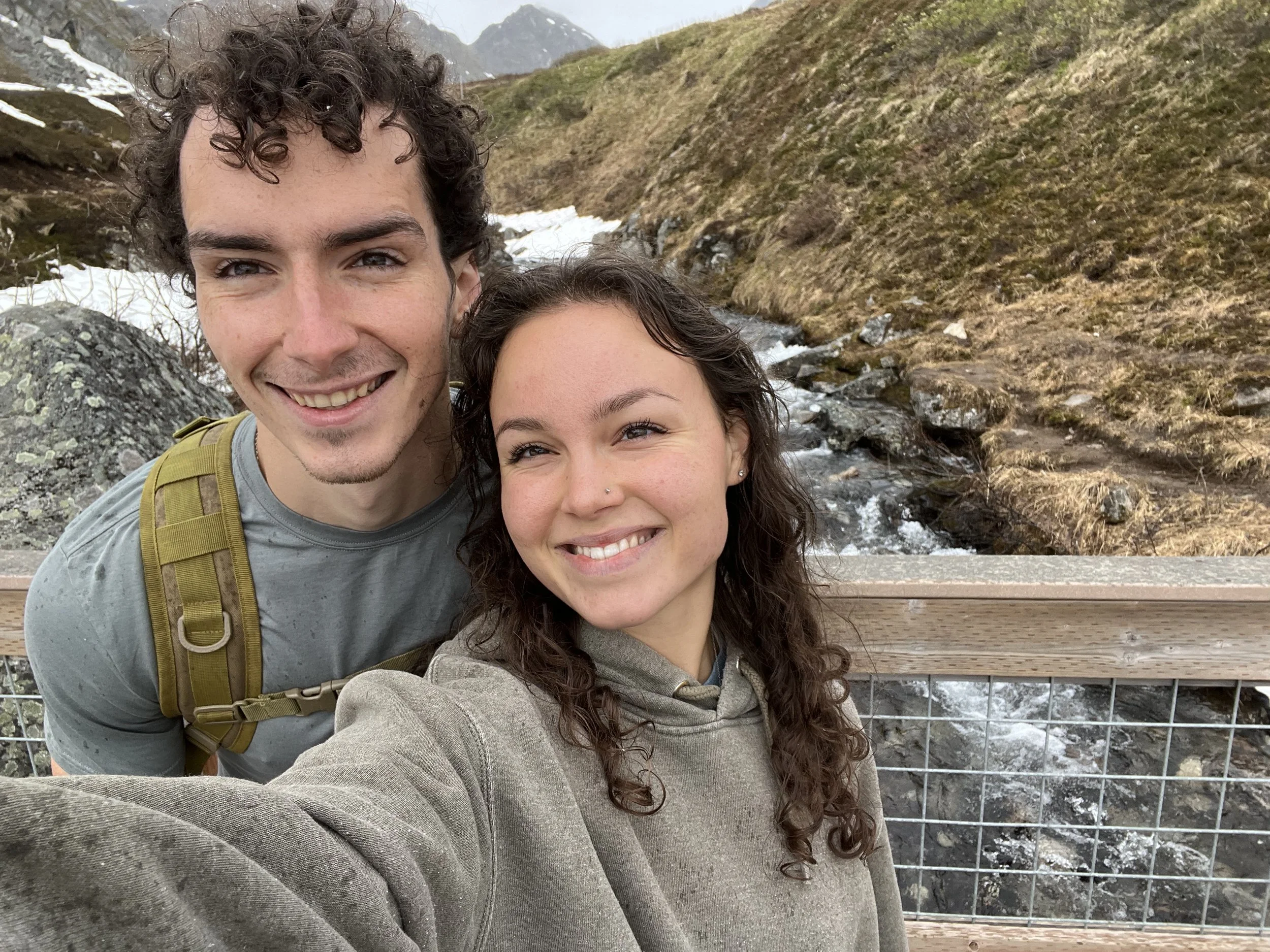 A smiling young man and woman taking a selfie on a wooden bridge overlooking a mountain stream. The background features rocky, snow-dusted terrain and distant mountains.