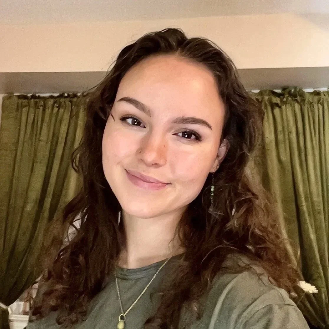 A young woman with brown curly hair, smiling softly, wearing a green top, earrings, a necklace, and standing in front of a green curtain.
