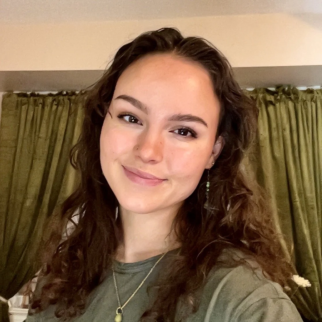 A young woman with long, curly brown hair, smiling, wearing a green top and a necklace, posing indoors with green curtains in the background.