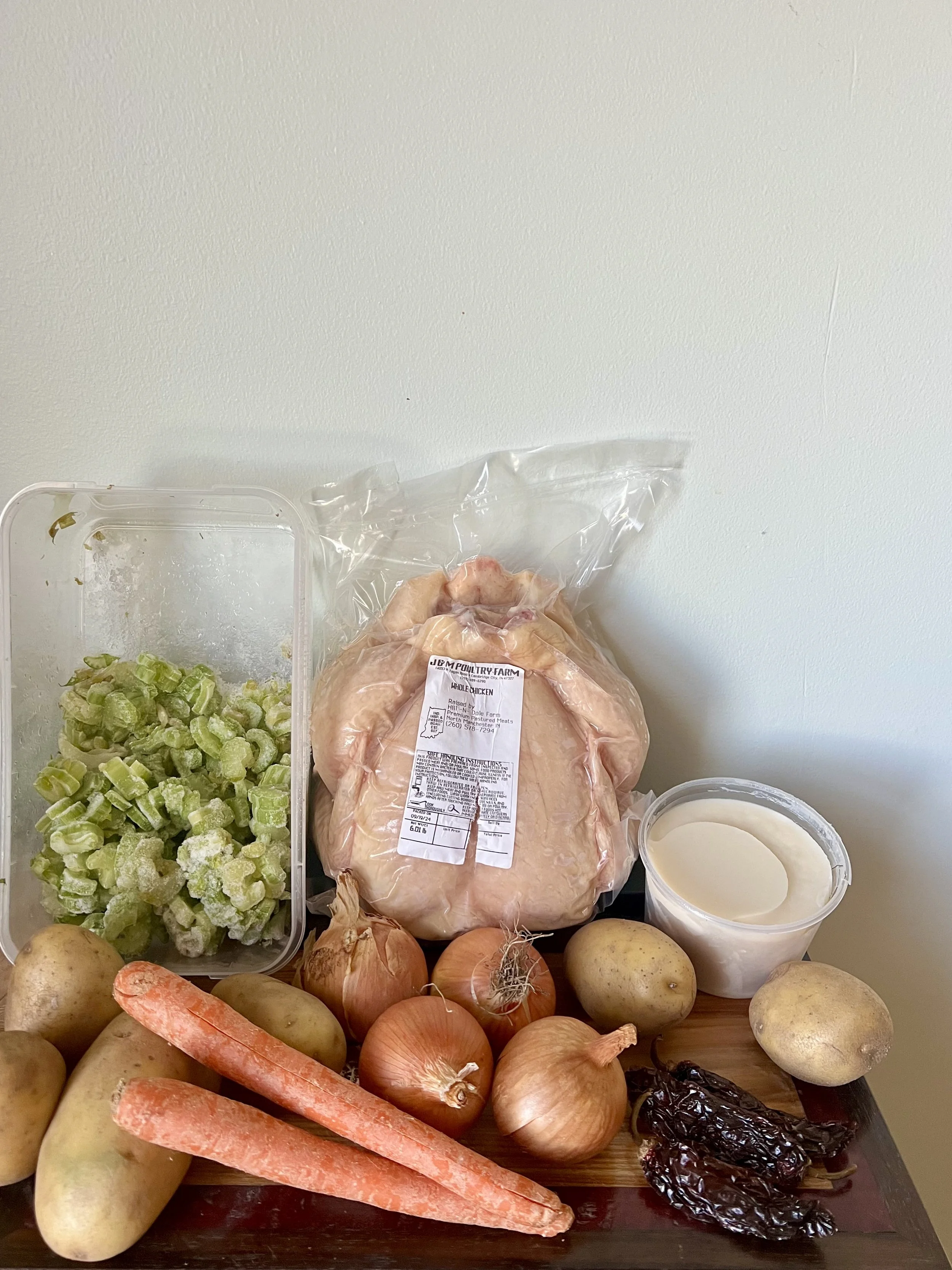 Raw whole chicken, potatoes, carrots, onions, poblano peppers, celery, and a container of sour cream on a wooden surface.