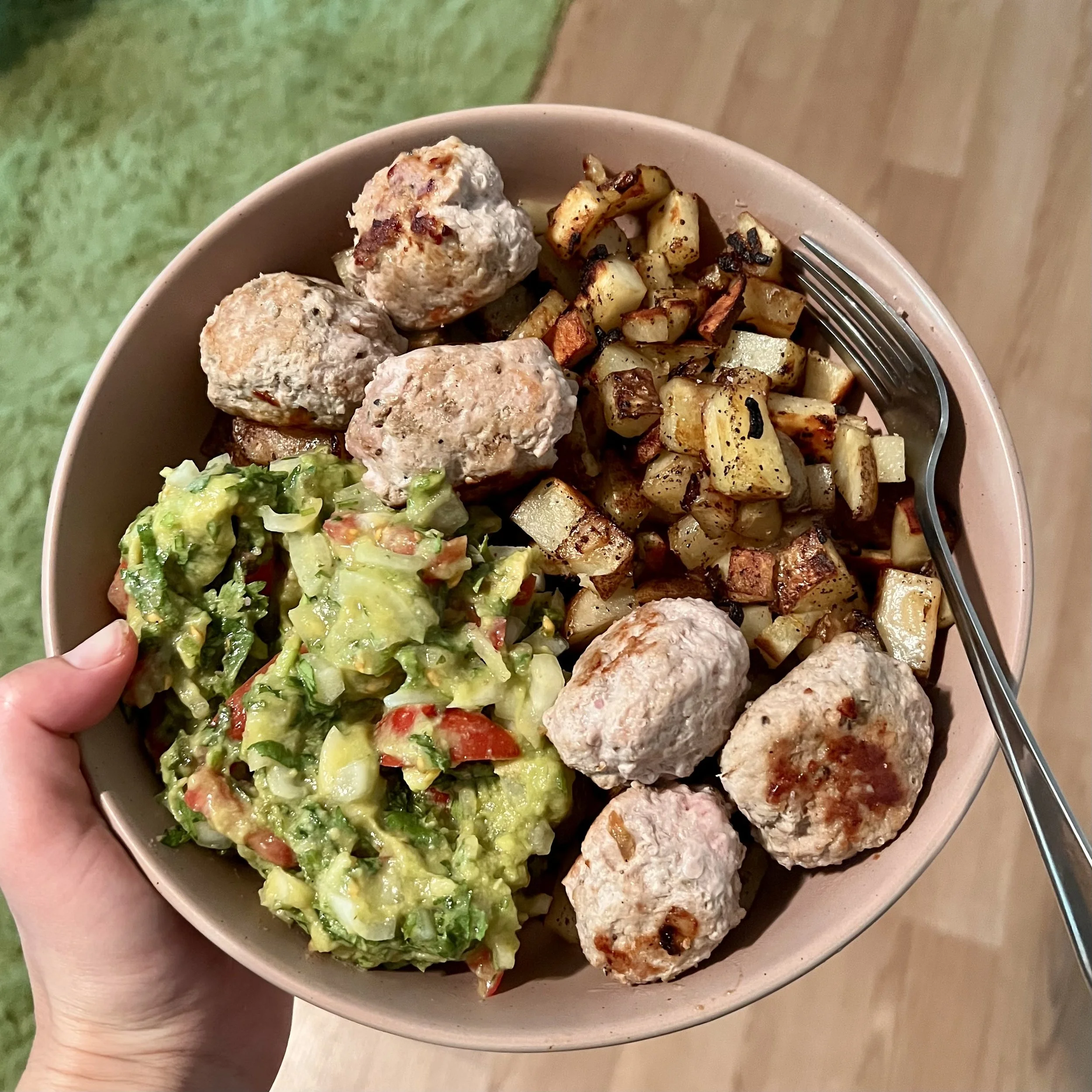 A bowl containing cooked meatballs, roasted diced potatoes, and guacamole with chopped tomatoes and cilantro.