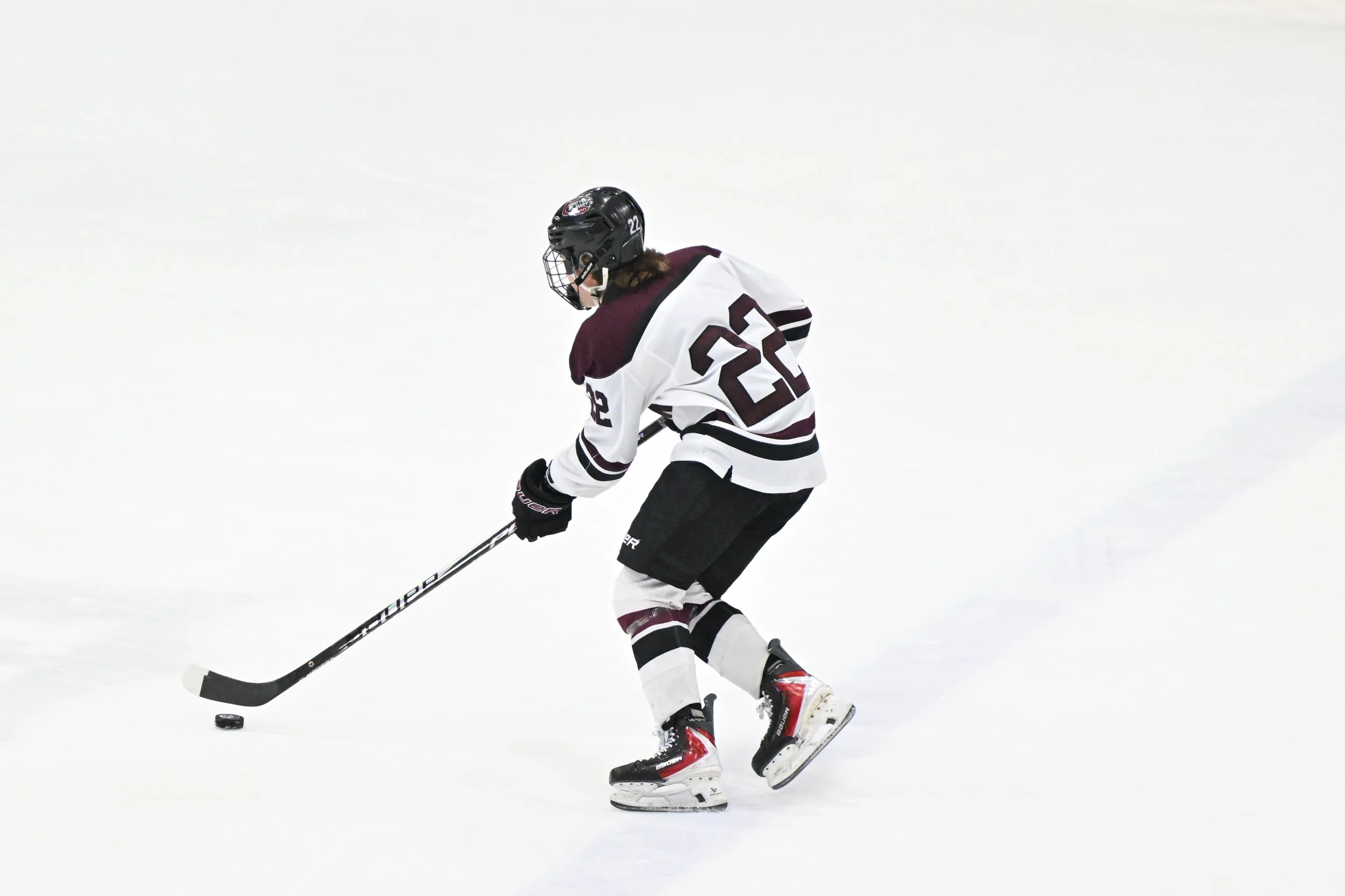 A hockey player in a white and maroon jersey with the number 22, wearing a black helmet, skating on ice and handling a hockey puck with a stick.