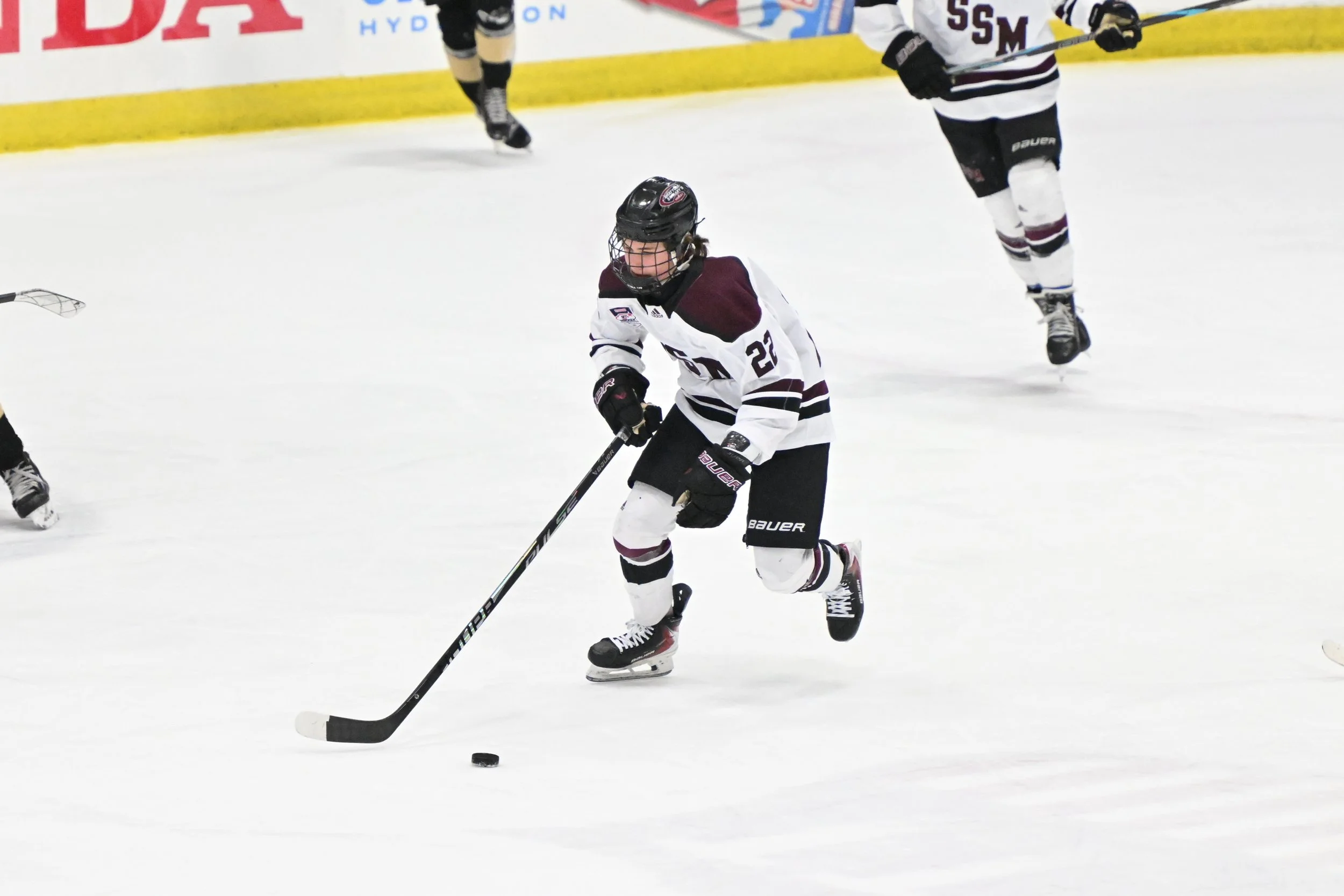 Hockey players on ice rink, one in foreground about to hit puck with stick, wearing white jersey with maroon and black details, black helmet, and skates. Other players are in background, one in black and white uniform, and the rink's yellow border an