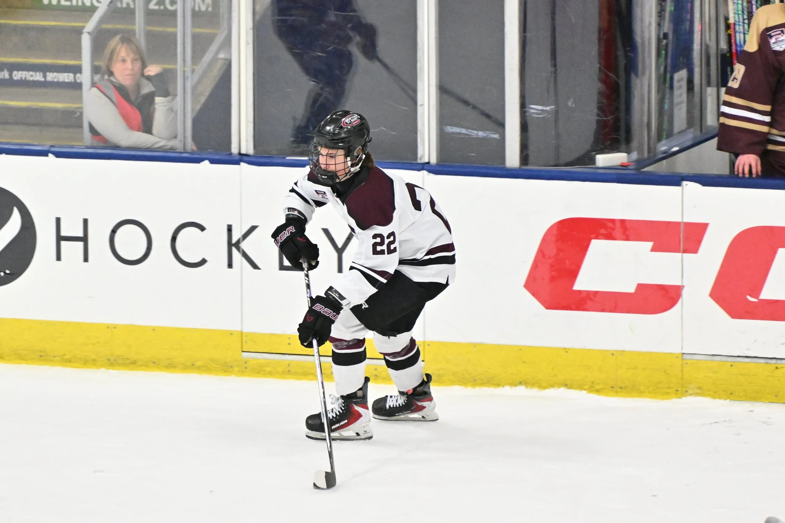 An ice hockey player wearing a black helmet, white and maroon jersey with the number 22, and black gloves, handling a hockey stick on the ice rink. Behind the player, a woman stands behind the glass, observing the game, with another person visible on