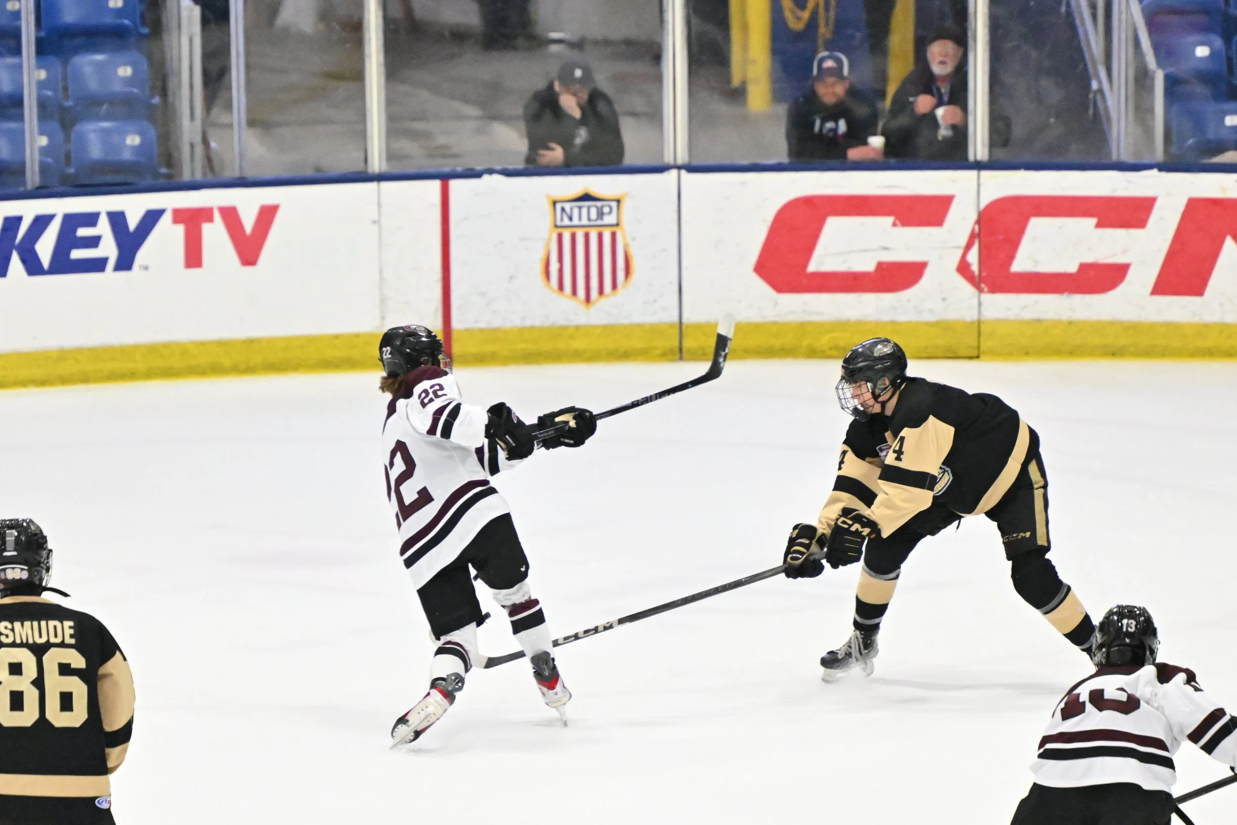 Hockey game in progress at an ice rink with three players fighting for the puck, spectators watching from behind the glass.