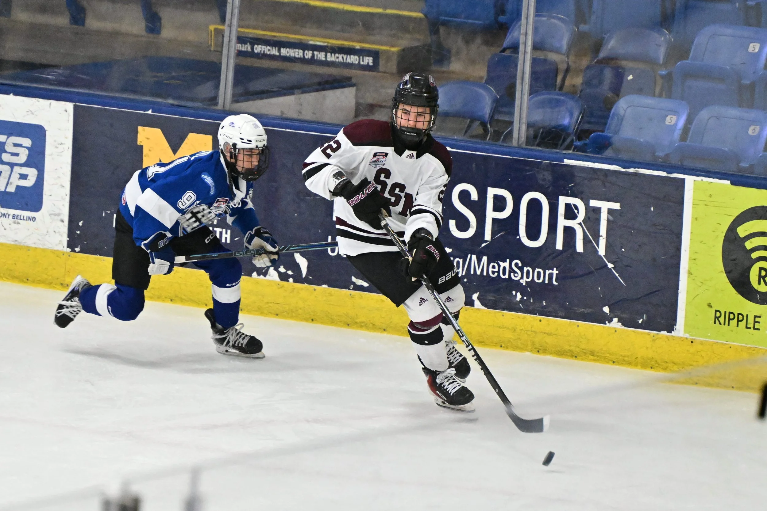 Two ice hockey players, one in a white jersey with maroon and black accents and the other in a blue jersey, are competing for the puck on the ice rink. The player in white is skating forward while the player in blue is attempting to reach the puck wi
