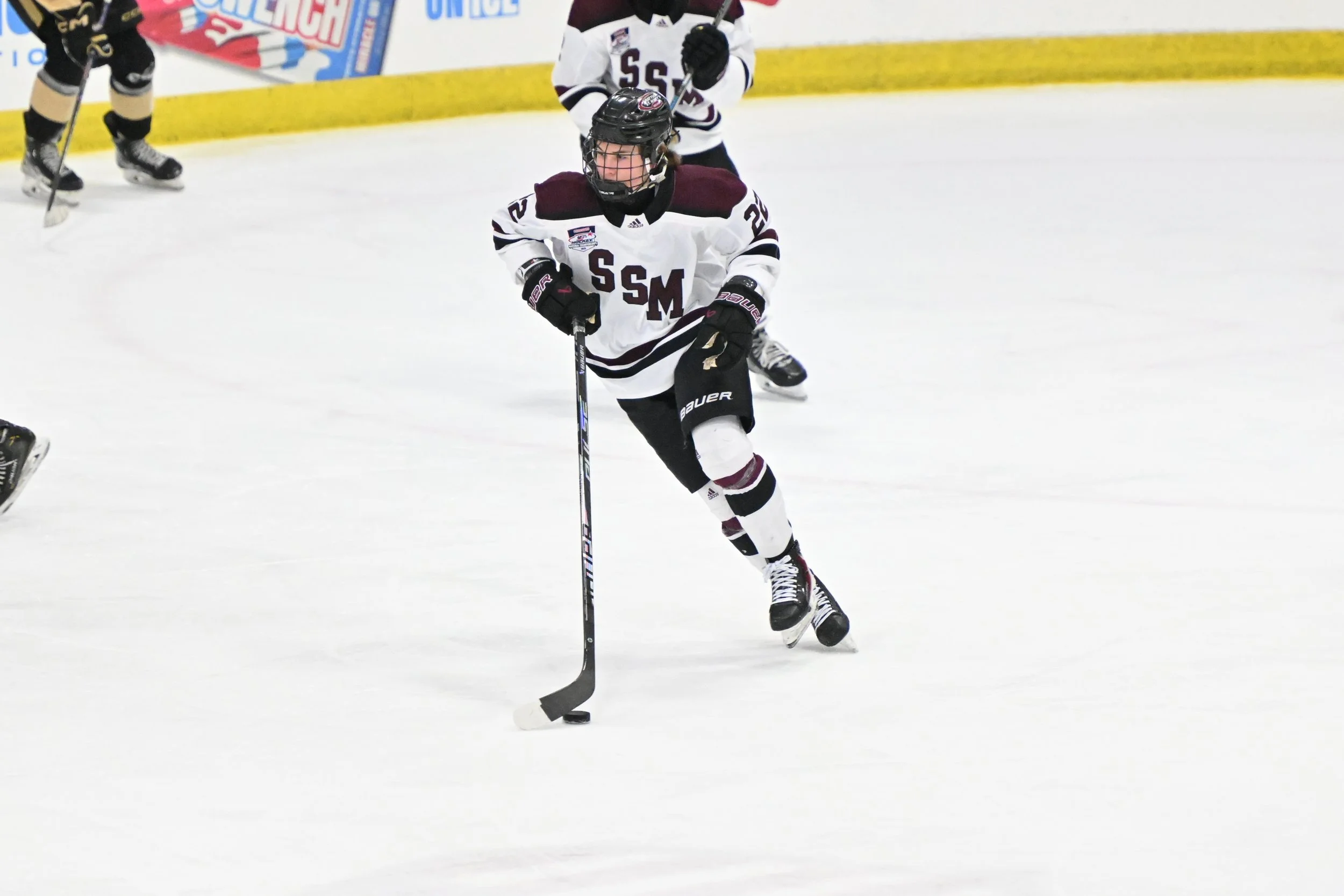 Hockey player in a white and maroon jersey skating on ice rink while holding hockey stick, with other players visible in the background.