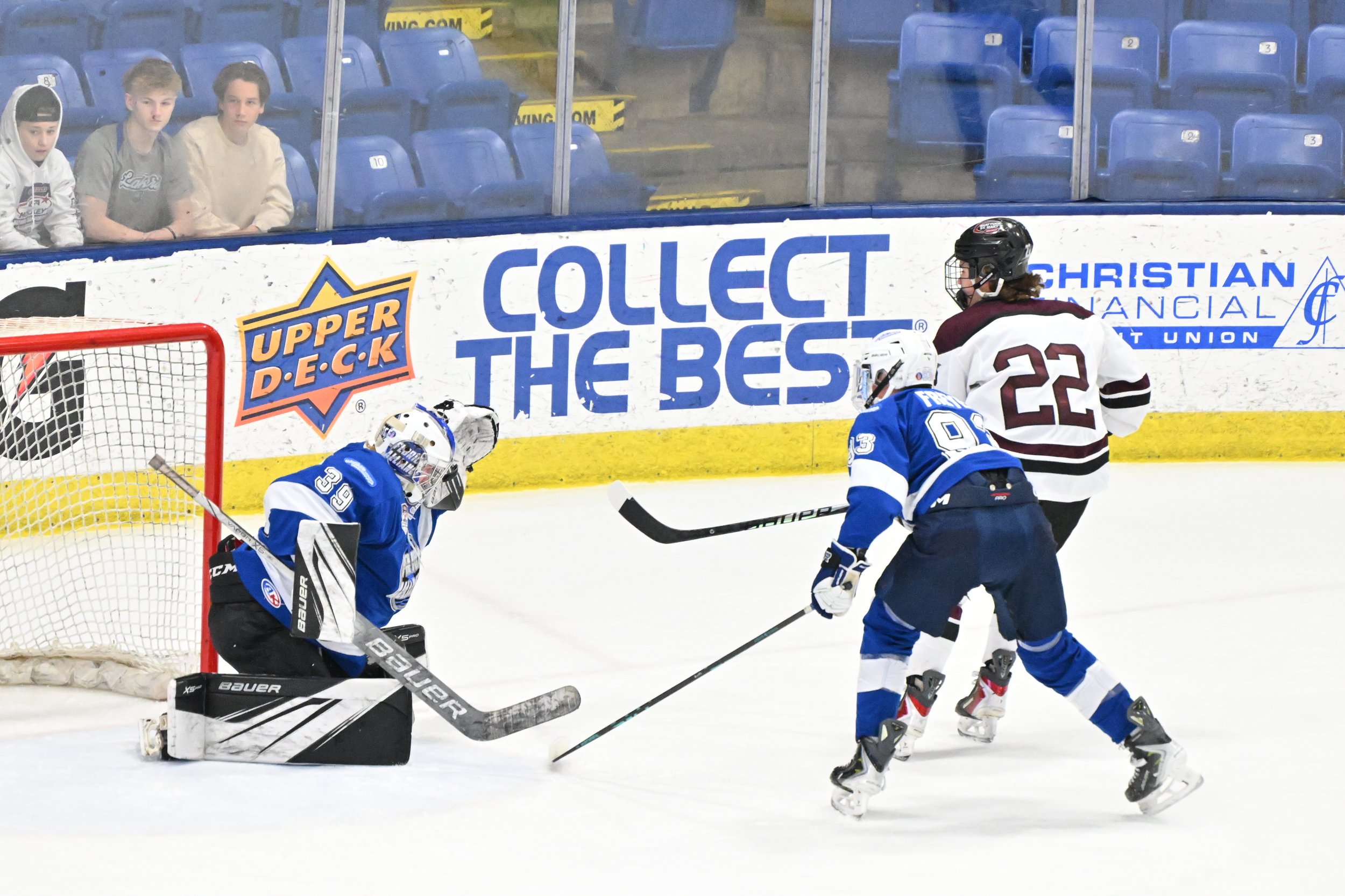 Ice hockey game in progress with three players, one goalie in blue gear, and an opposing player in a white jersey near the goal. Three spectators are watching from behind the glass, sitting in blue seats.