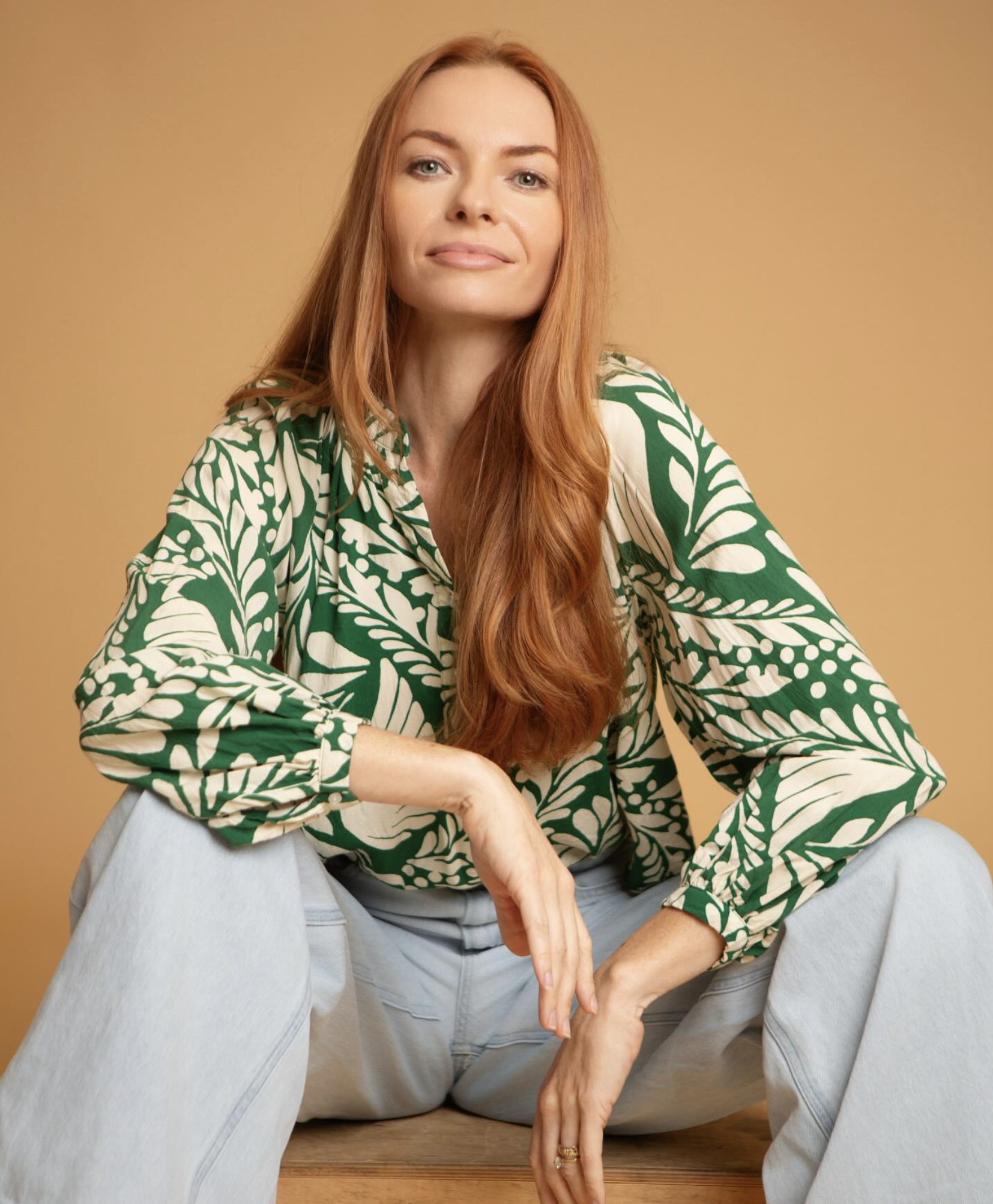 A woman with long red hair wearing a green and cream patterned blouse and light-colored pants, sitting on a wooden surface against a tan background.
