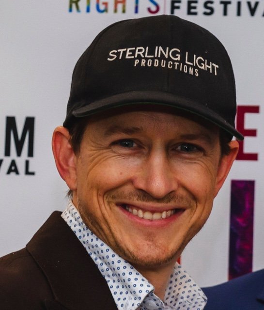 A smiling man wearing a black baseball cap with the text 'Sterling Light Productions' and a white shirt with small blue polka dots, standing in front of a backdrop at an event.