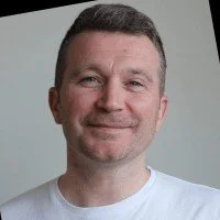 A headshot of a man with short brown hair, a light beard, and a slight smile, wearing a white shirt against a neutral background.