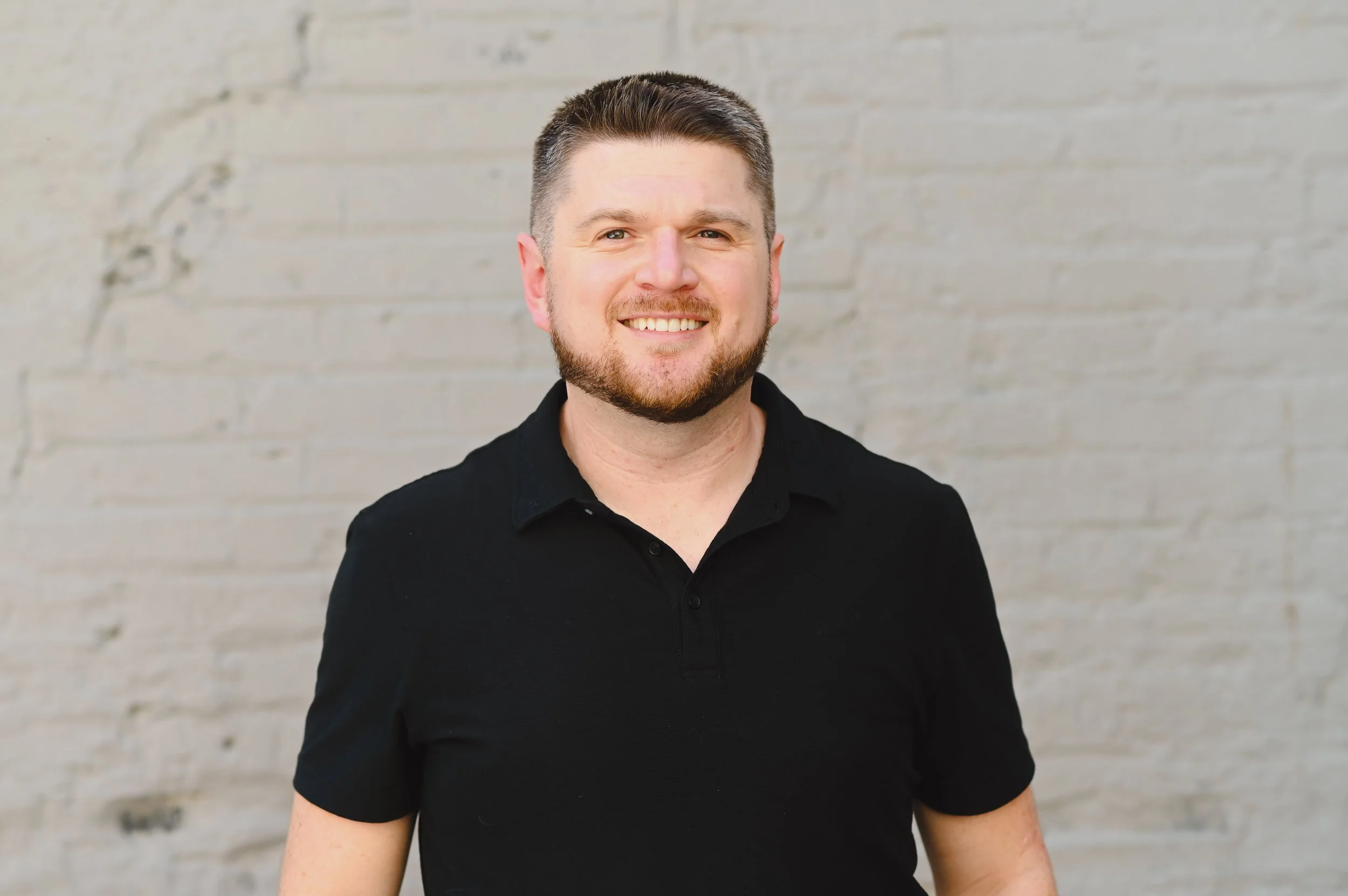 A smiling man with short dark hair and a beard, wearing a black collared shirt, standing in front of a light gray brick wall.