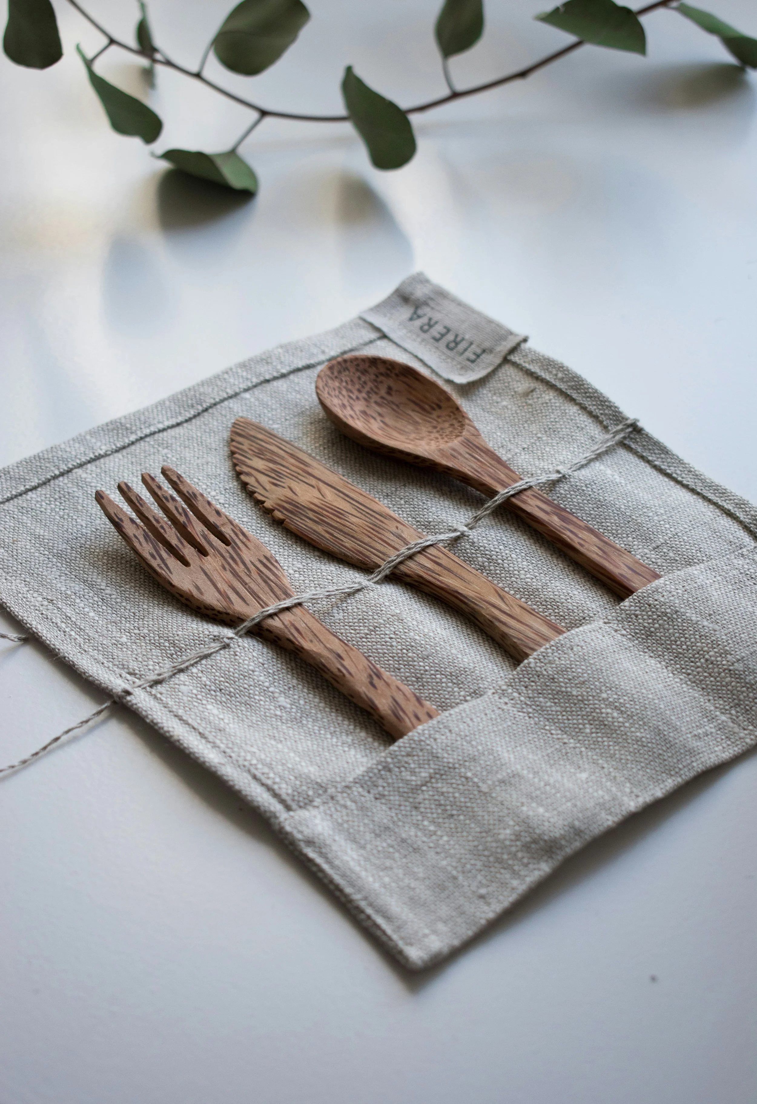 Wooden fork, knife, and spoon on a linen holder with the word 'FIRER'A' visible, on a white surface, with green leaves in the background.