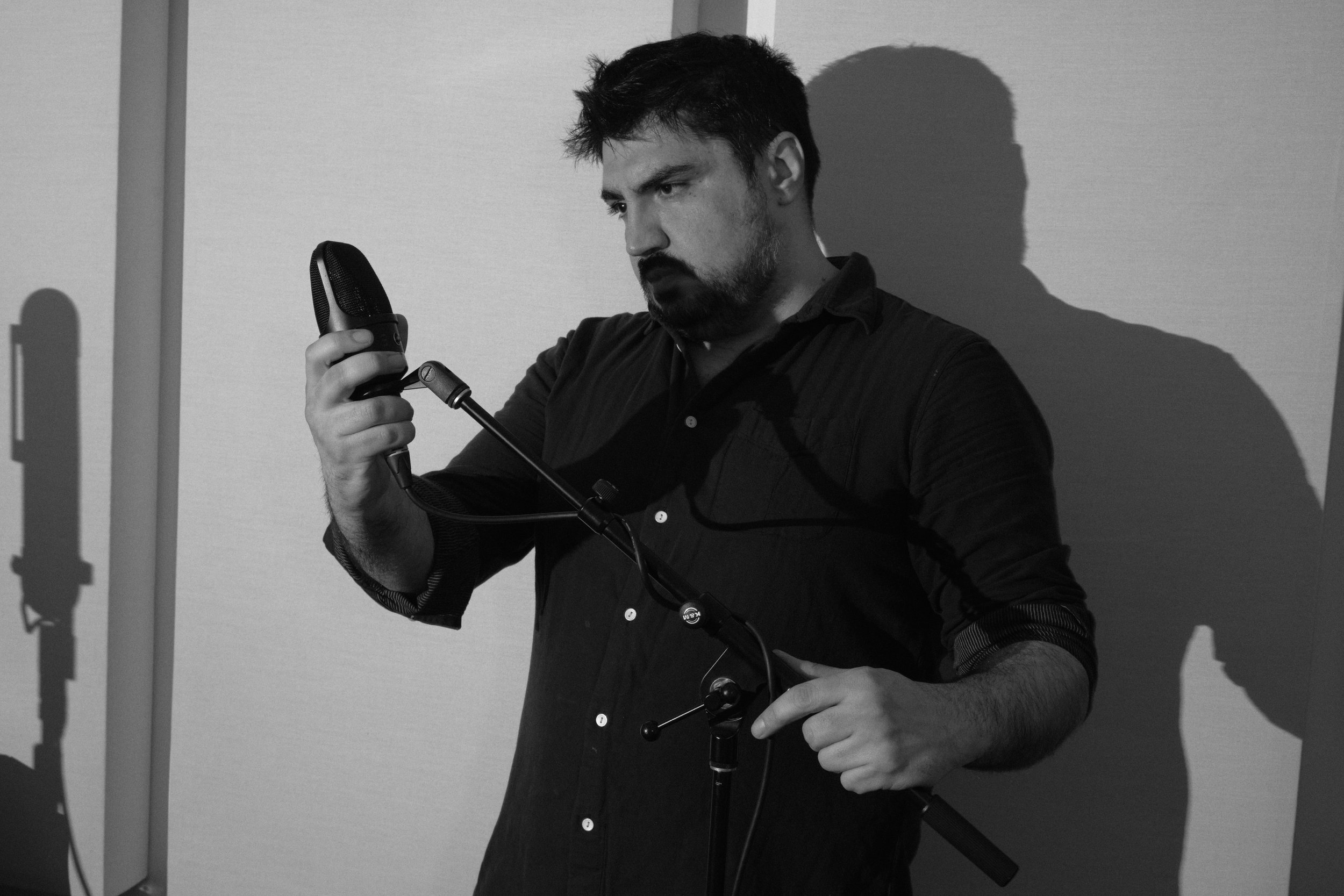 A man with dark hair and a beard, dressed in a dark shirt, holding a professional microphone and a microphone stand, standing against a plain wall with his shadow cast behind him.