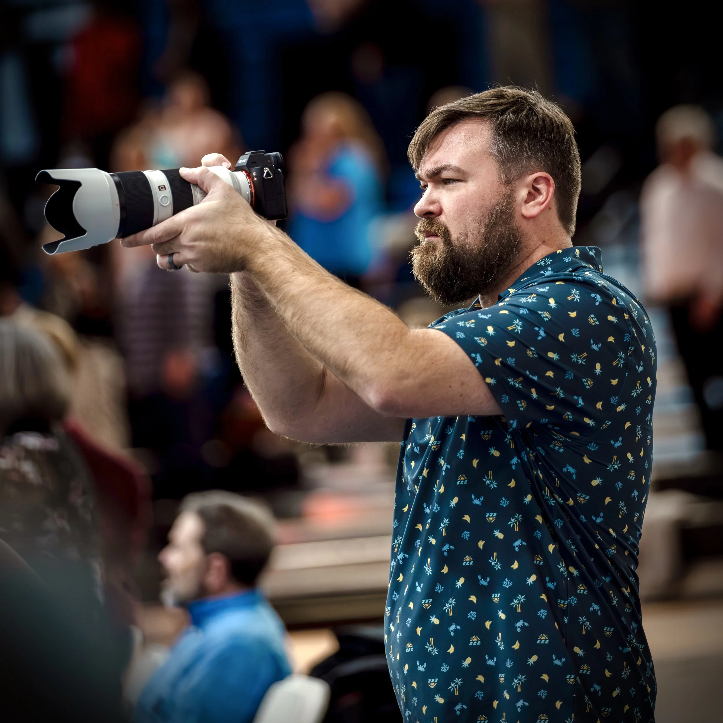Stephen wearing a blue shirt with a tropical pattern, is holding a professional camera with a large telephoto lens and taking a picture in a crowd. The background is blurred with other people present.