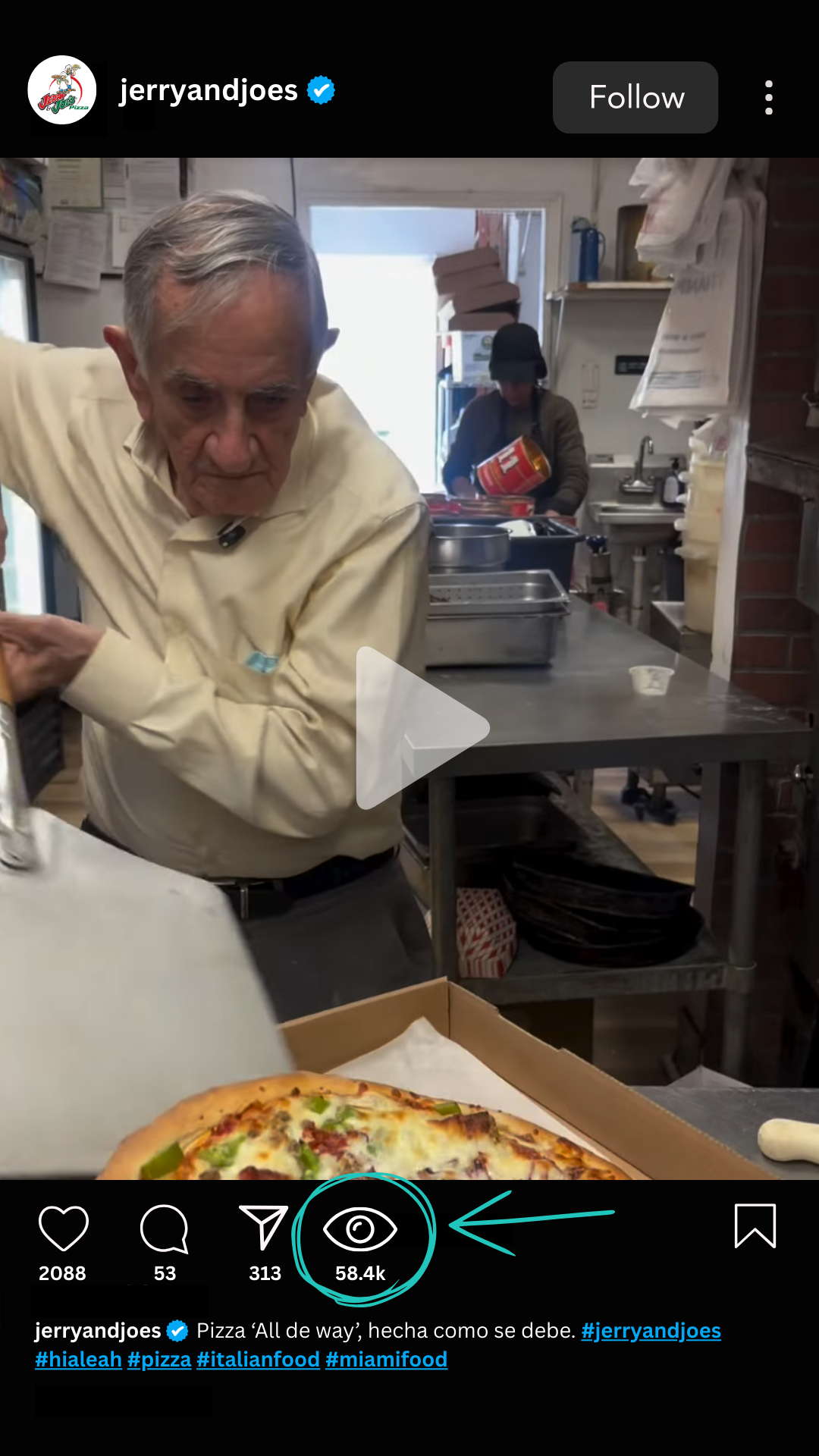 Old man with gray hair in a maroon shirt standing in a pizzeria kitchen, smiling at the camera.