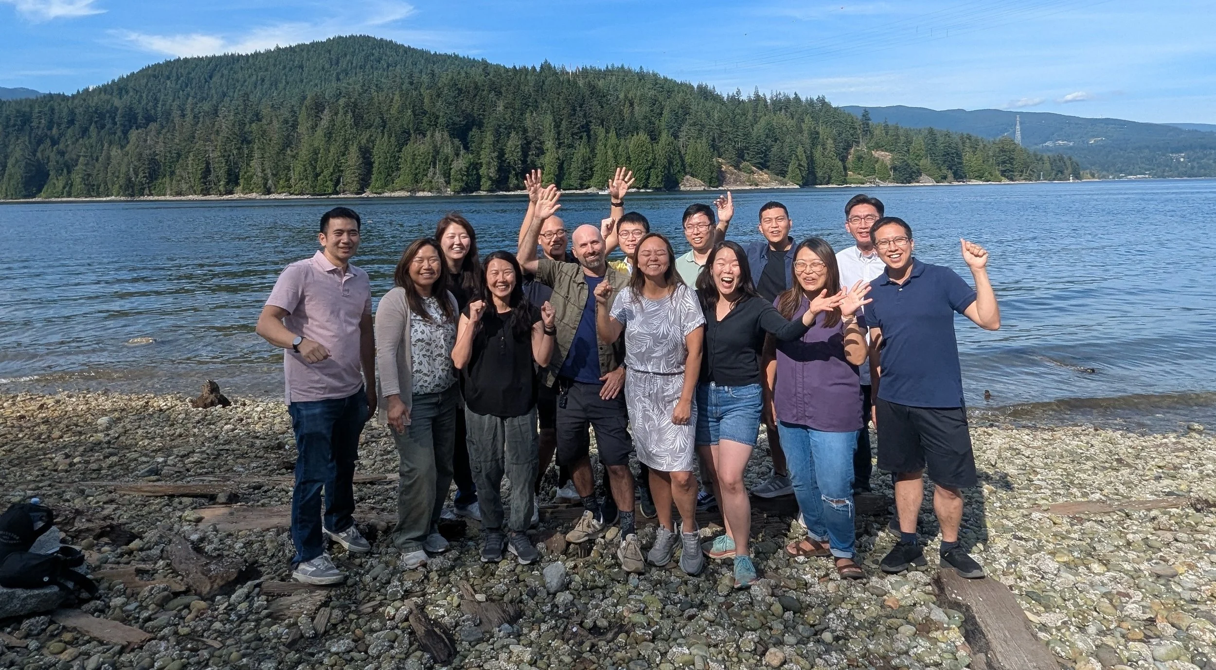 Group of people standing on a rocky lakeshore with trees and mountains in the background, smiling and celebrating.