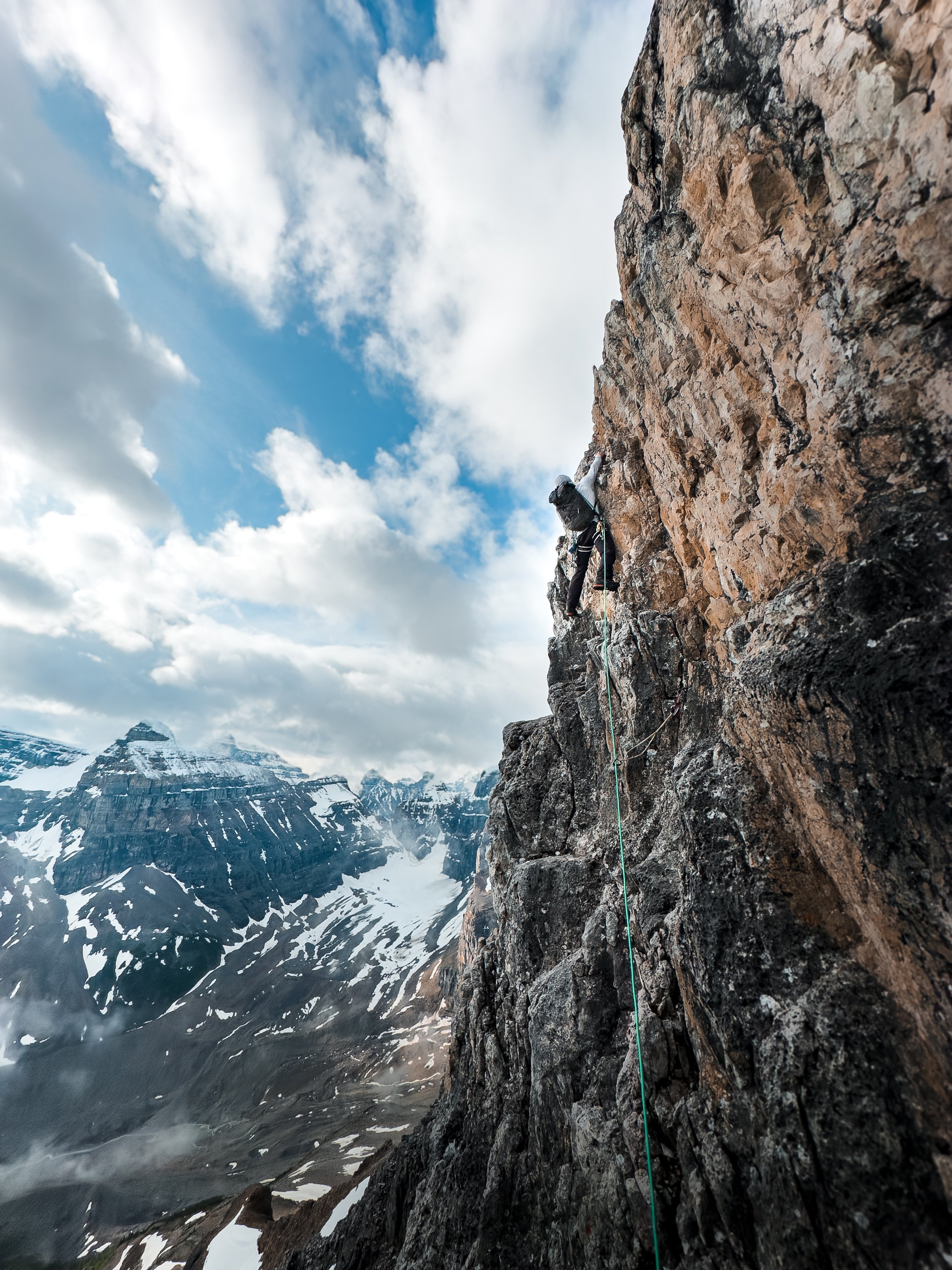 A man with a black backpack climbing a steep rock face with snowy mountains and glaciers in the distance