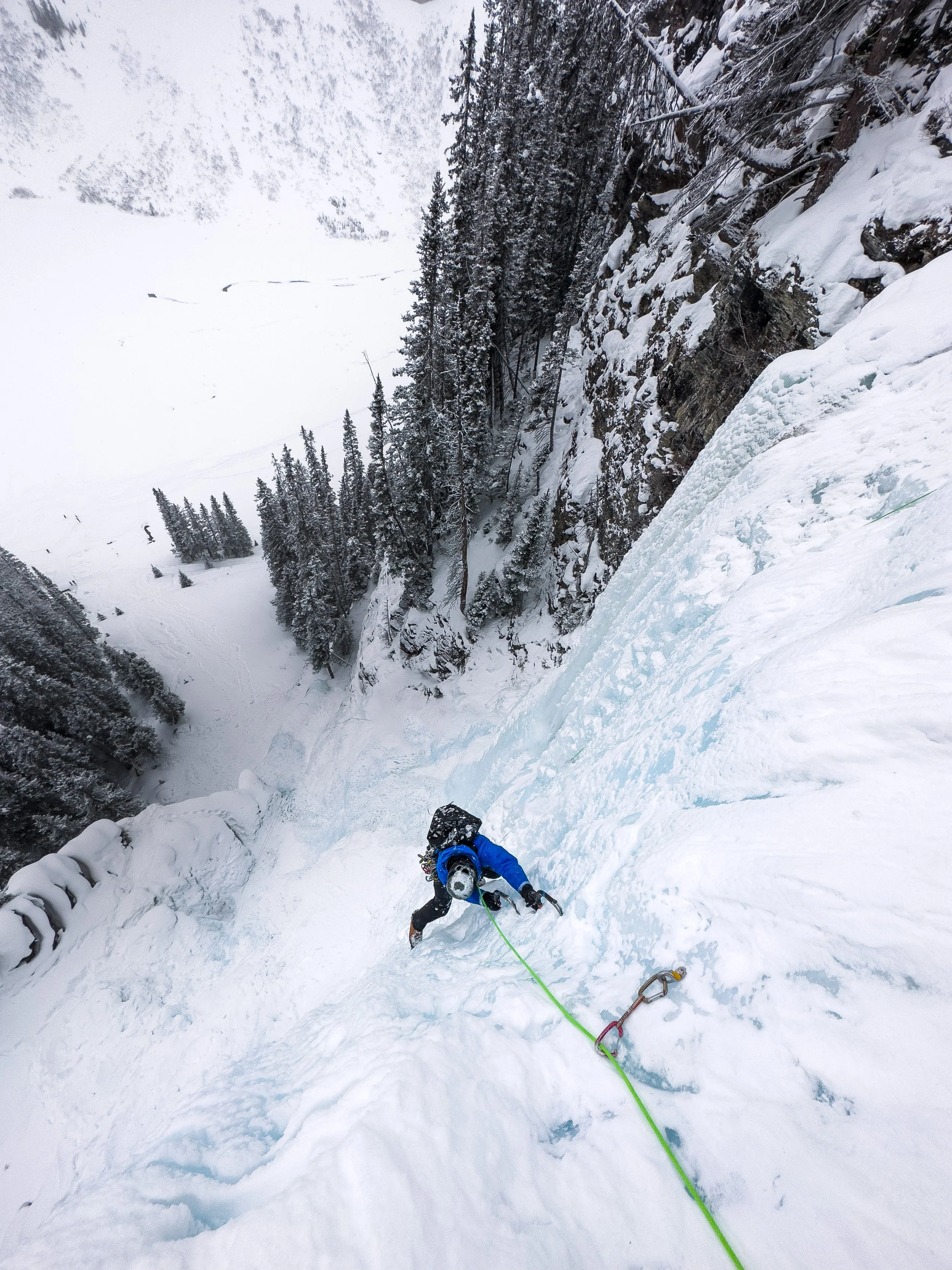A person ice climbing on a frozen waterfall in a snowy mountain landscape, wearing blue and black gear with a green climbing rope.