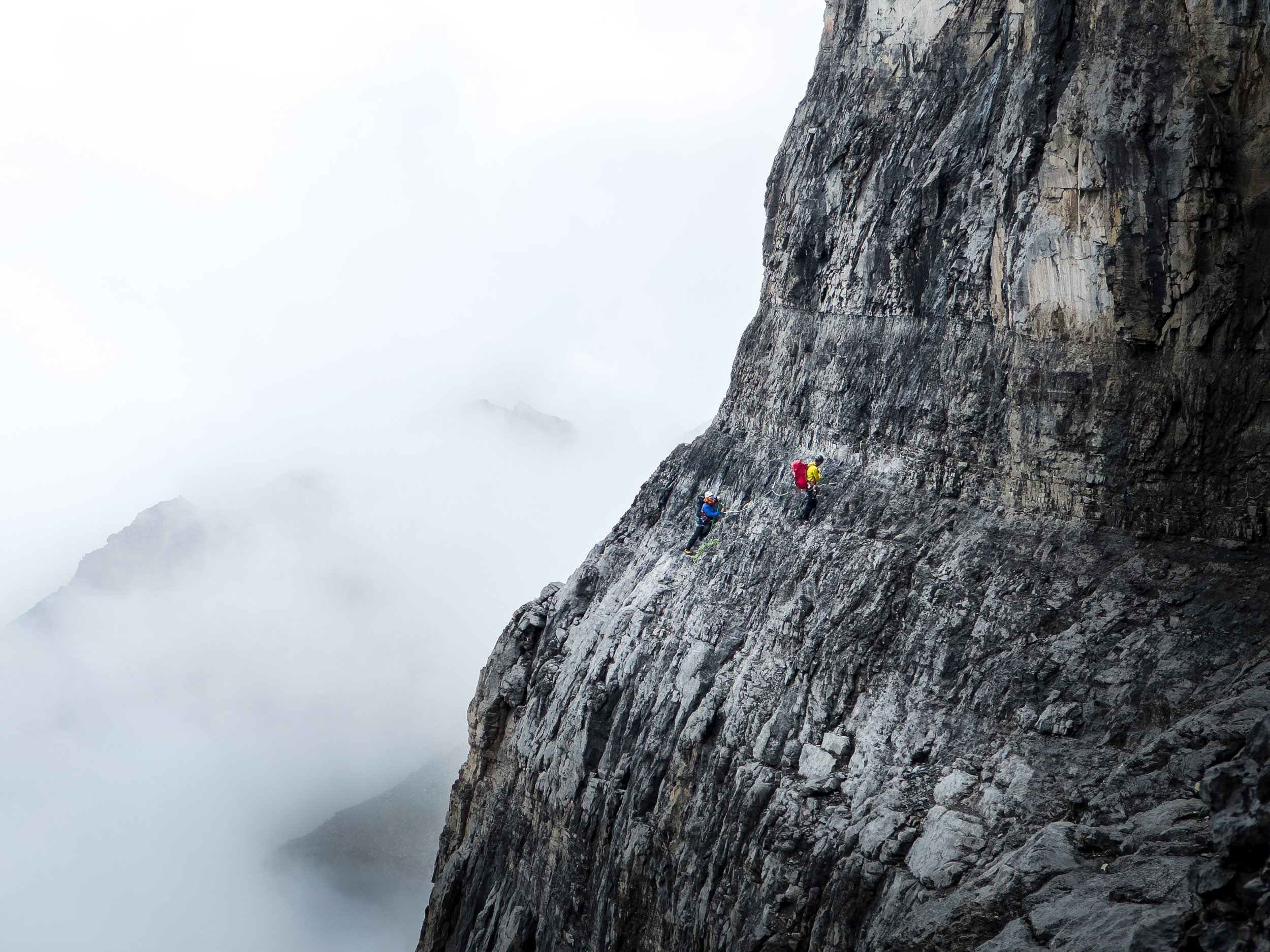 Two rock climbers ascending a steep, rugged mountain face with clouds and mist surrounding them.