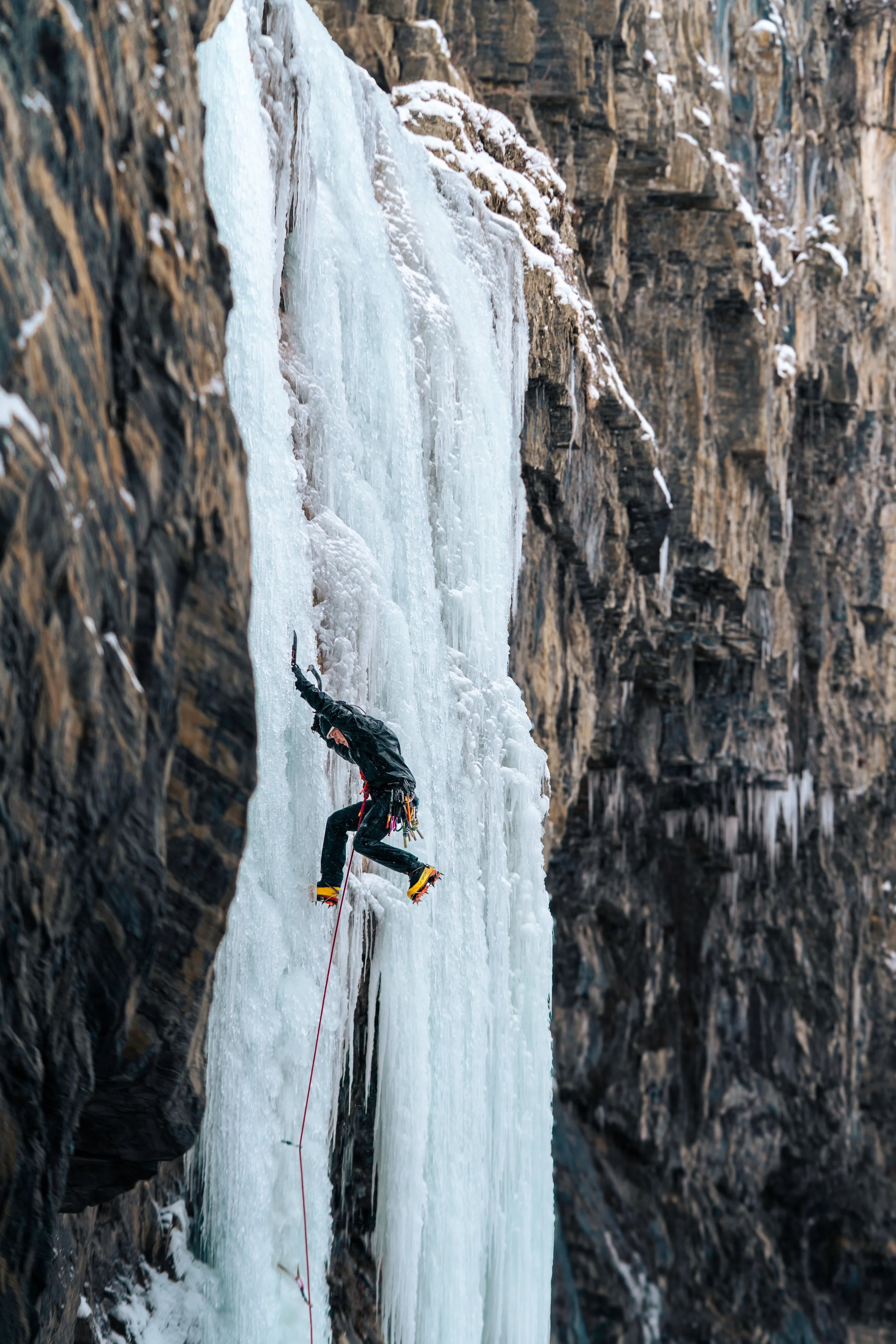 A man in a black jacket and yellow boots ice climbing on a steep frozen waterfall